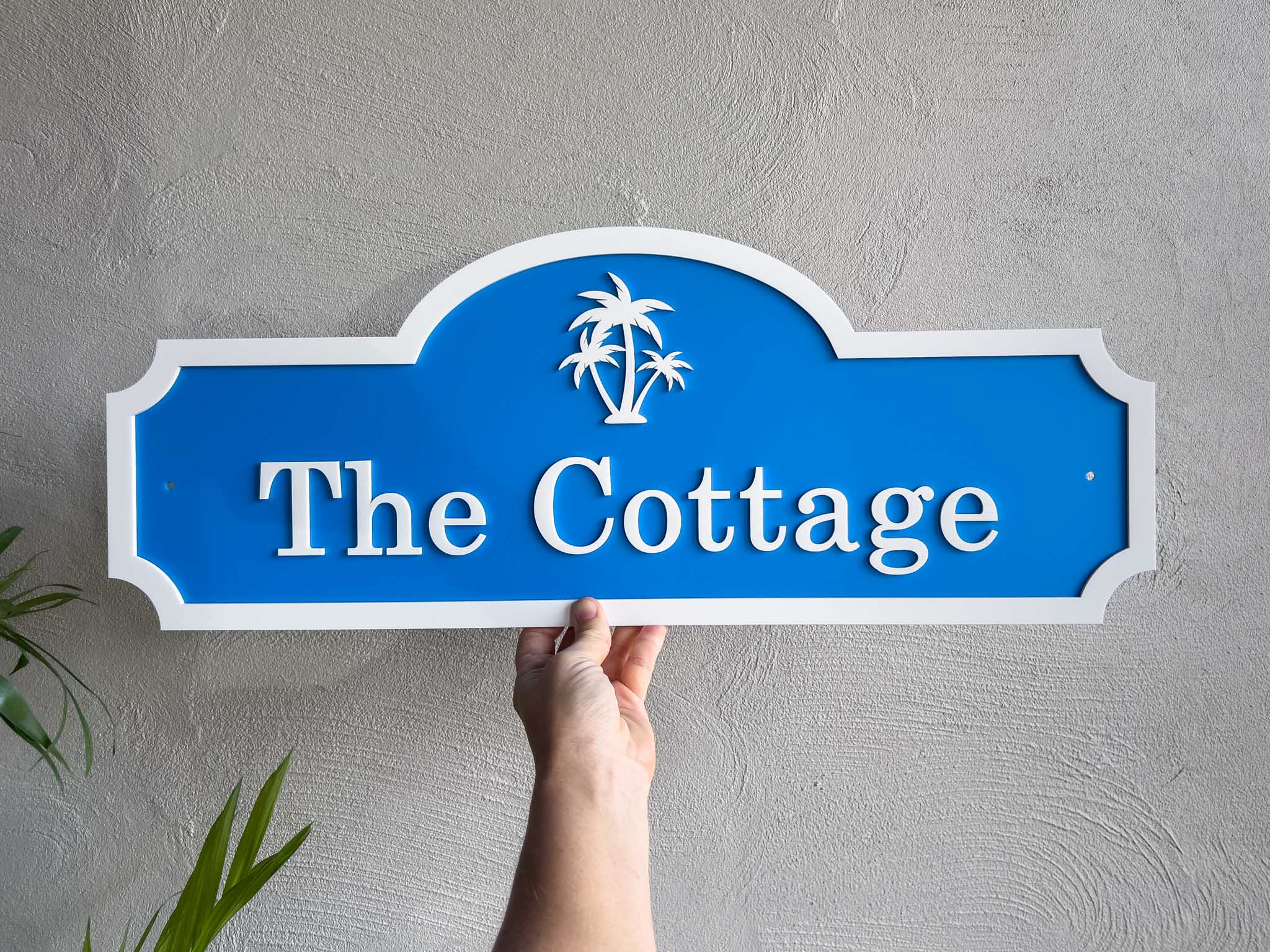 Hand holding a blue sign with 'The Cottage' and palm tree design against a gray wall.