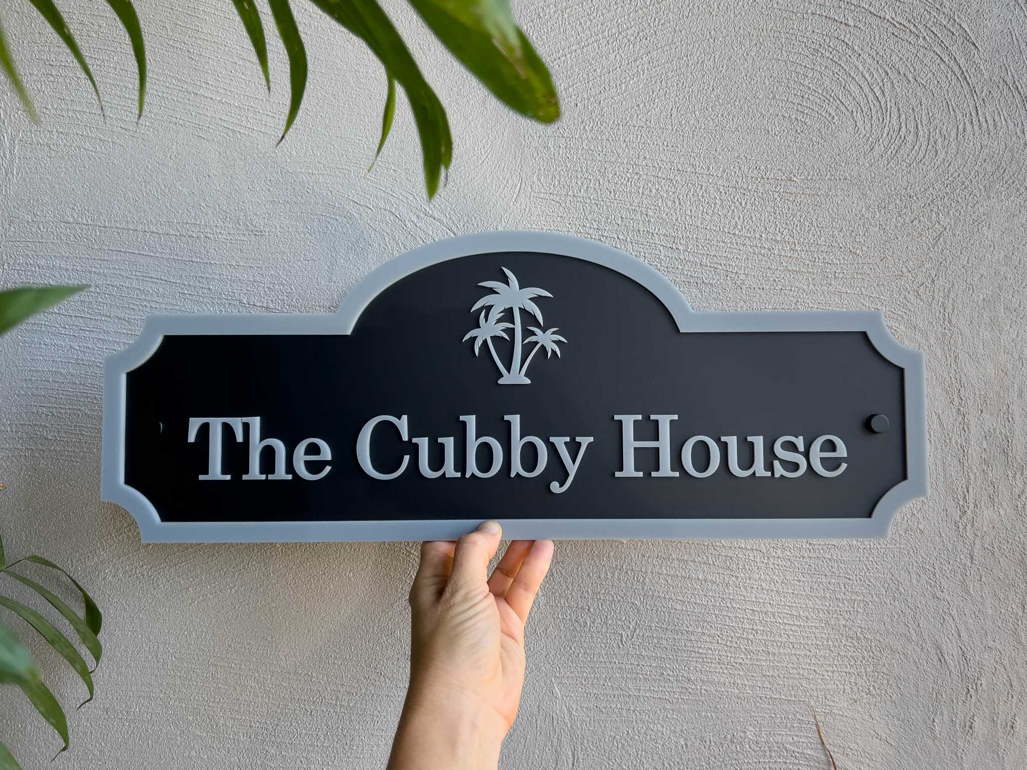 Hand holding a personalised black and grey sign with 'The Cubby House' text against a light grey wall.