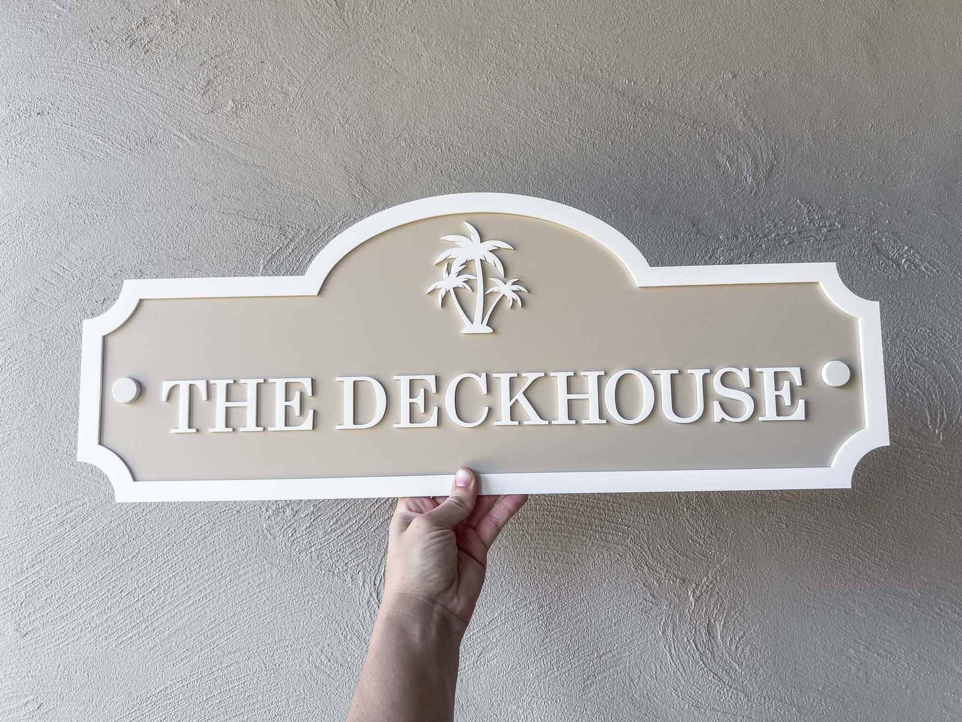 Hand holding a 3D acrylic decorative sign with 'The Deckhouse' text against a plain wall.