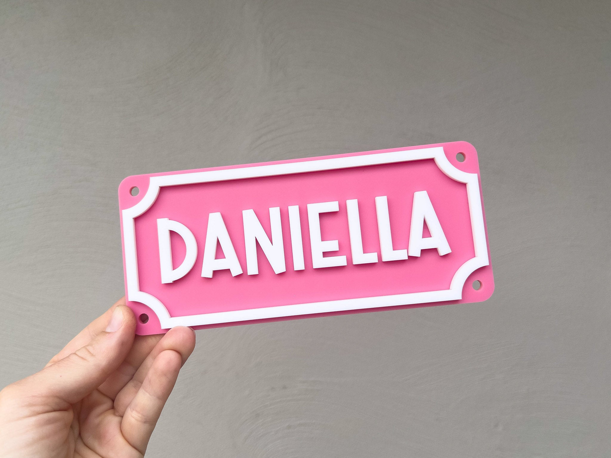 Pink nameplate with 'DANIELLA' held by a hand against a gray background