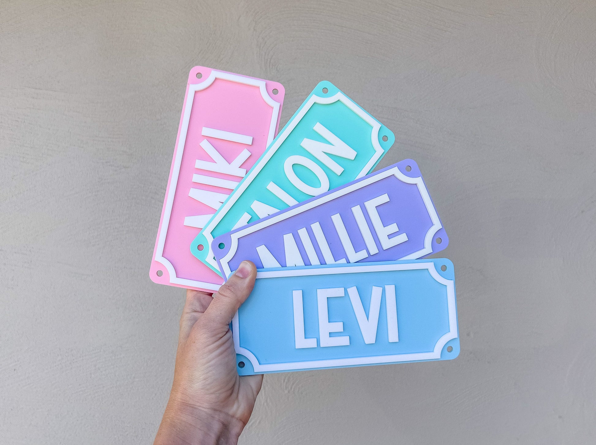 Hand holding four colorful name plates with names 'Miki', 'Talon', 'Millie', and 'Levi' against a plain background.