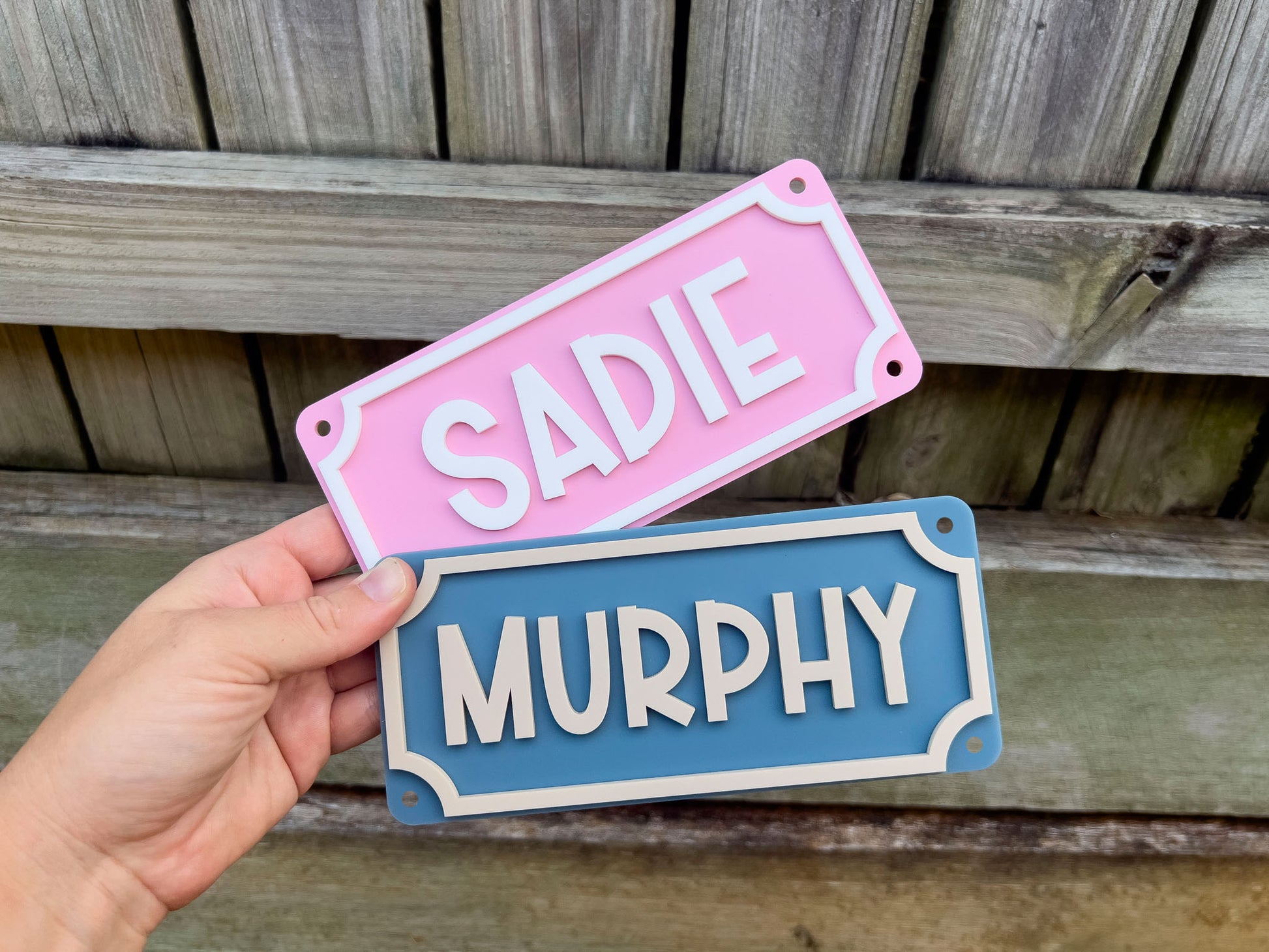 Two nameplates, one pink and one blue, held by a hand against a wooden background.