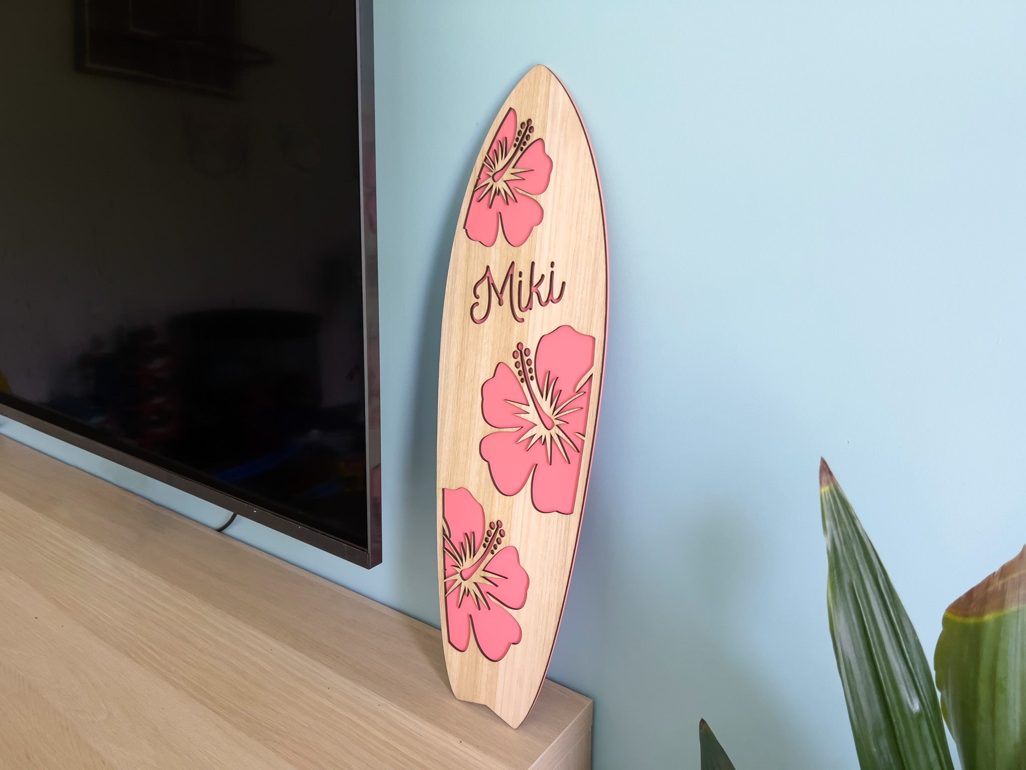 Wooden surfboard with a salmon pink hibiscus flower design and the name 'Miki' against a light blue wall.