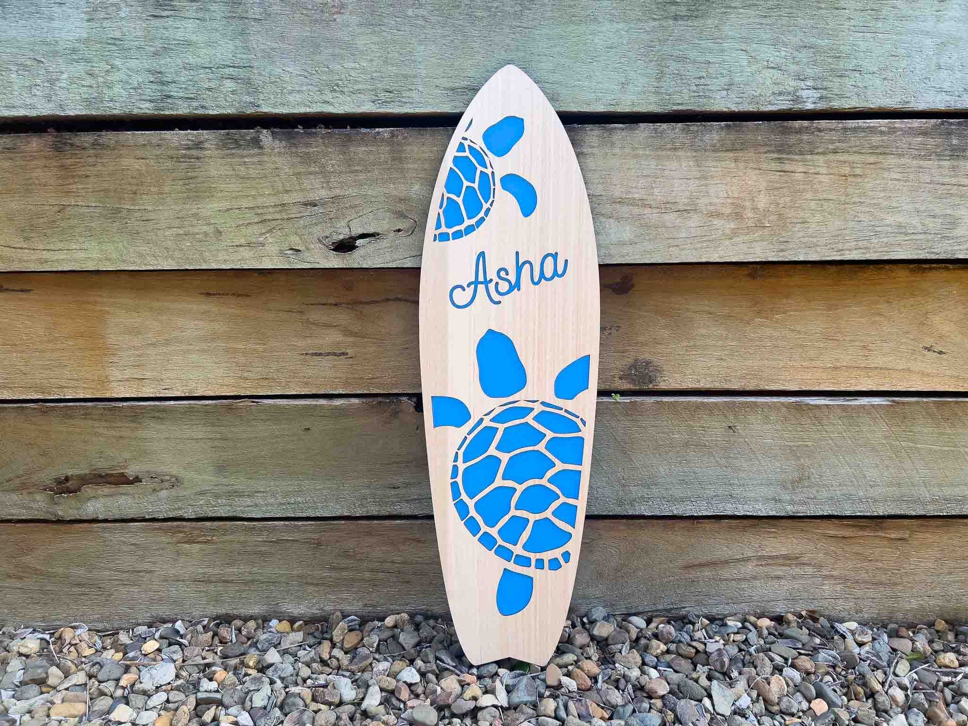 Wooden surfboard with blue turtle design and 'Asha' branding against a wooden wall.