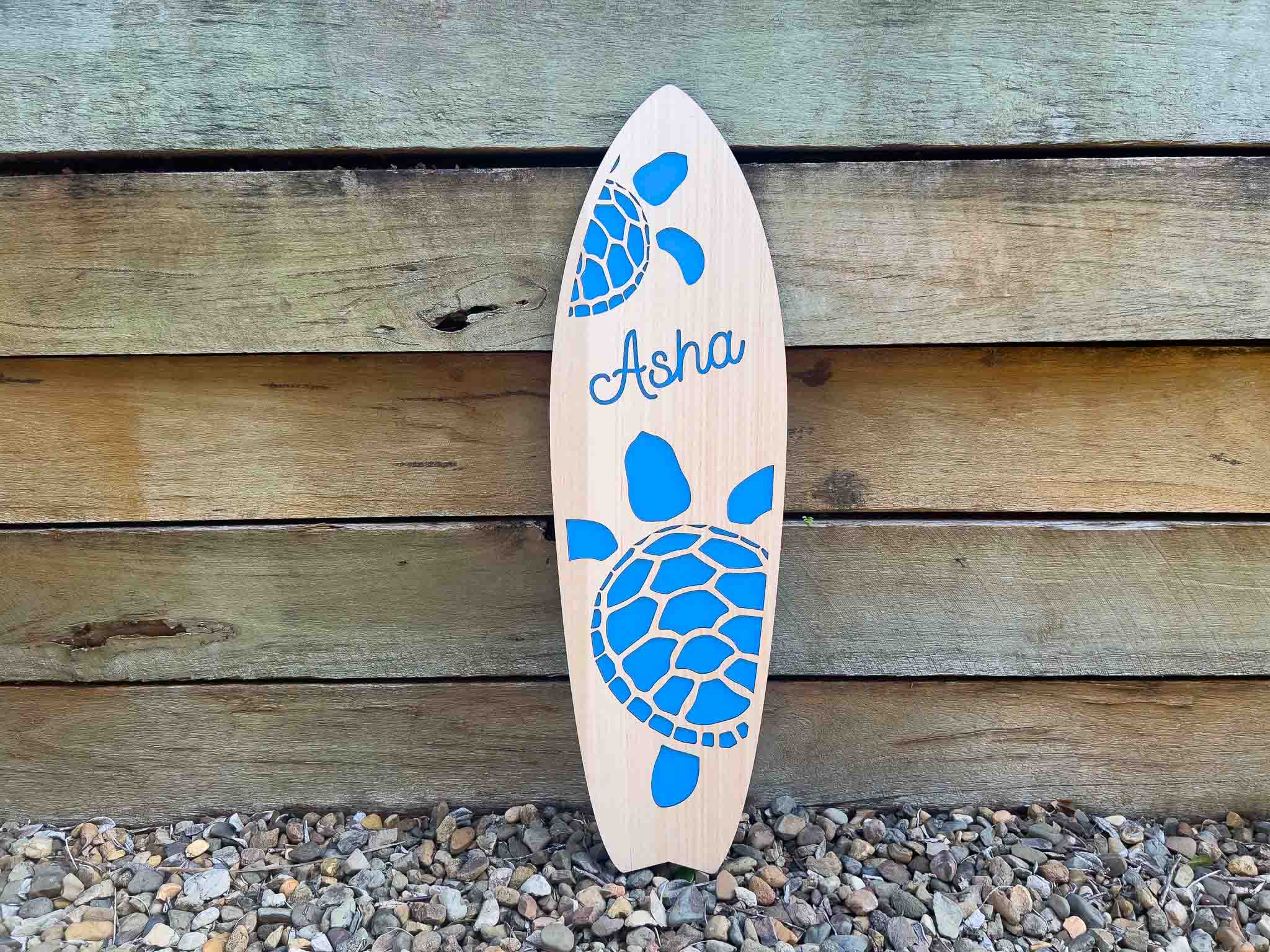 Wooden surfboard with blue turtle design and 'Asha' branding against a wooden wall.