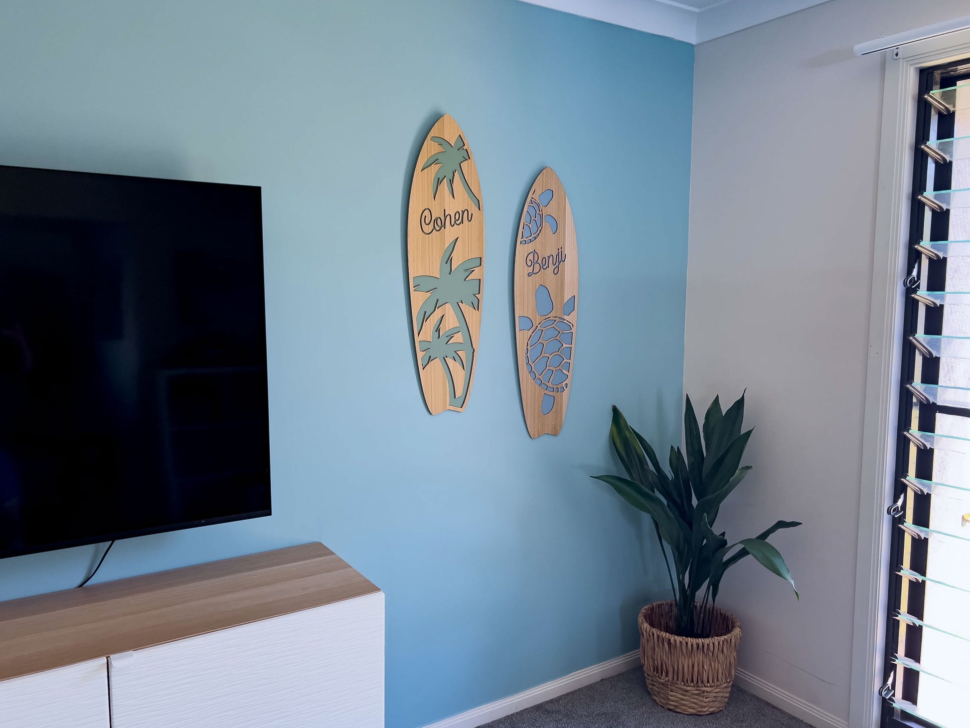 Two timber surfboard name plaques mounted to a wall in a coastal aesthetic living room.