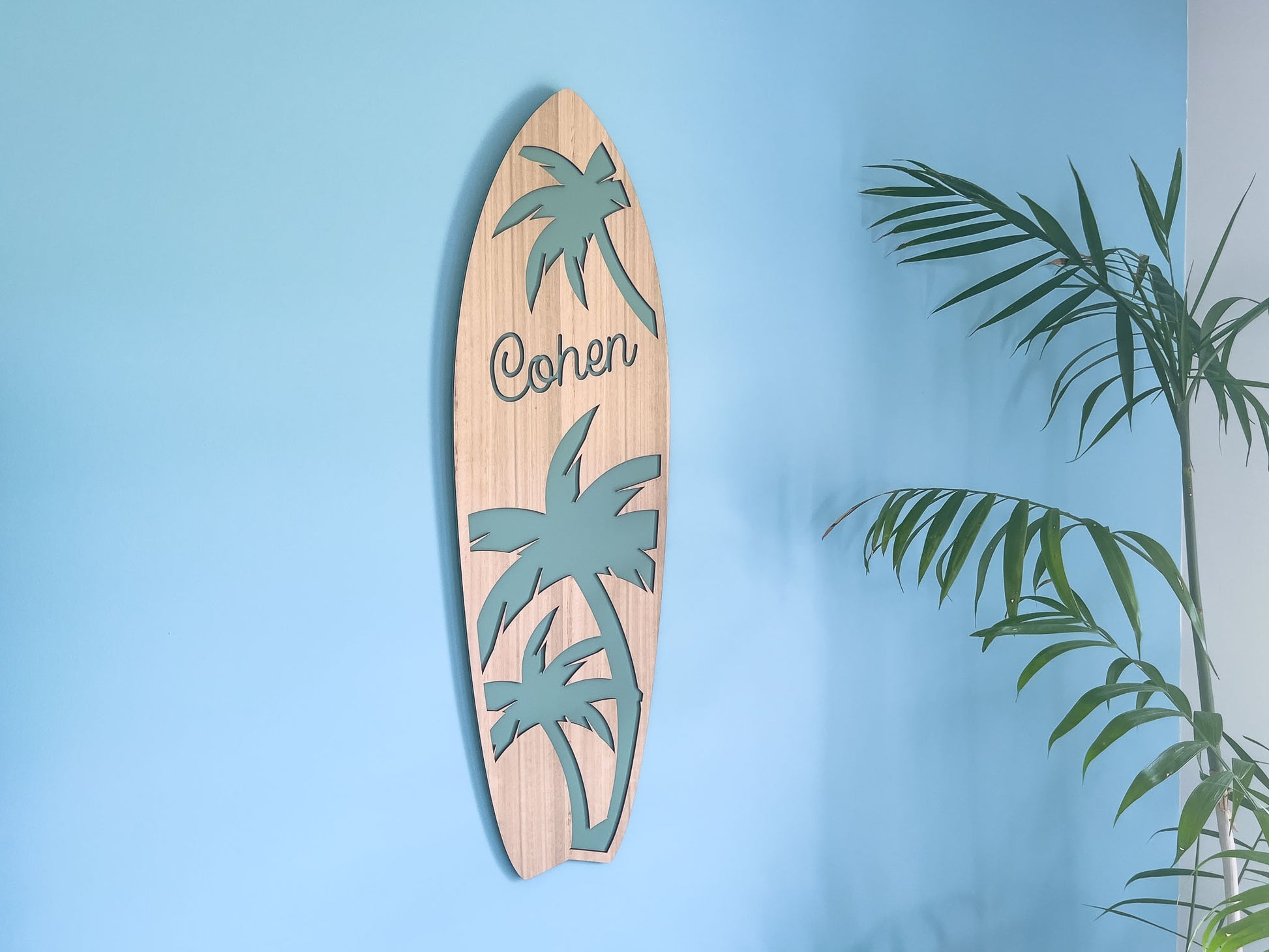 Wooden surfboard-shaped wall decoration with palm tree designs and the name 'Cohen' on a blue wall.