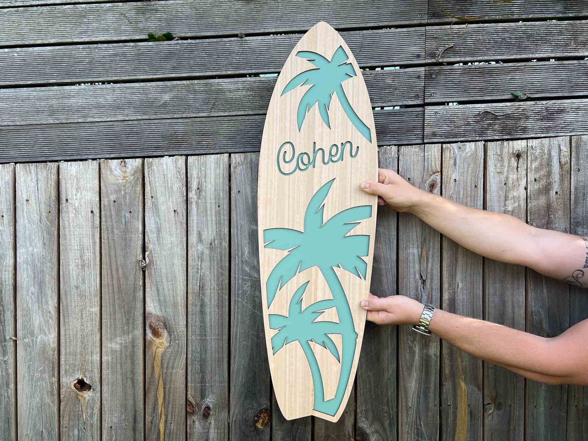 Wooden surfboard-shaped sign with palm tree designs and the name 'Cohen' against a wooden background.
