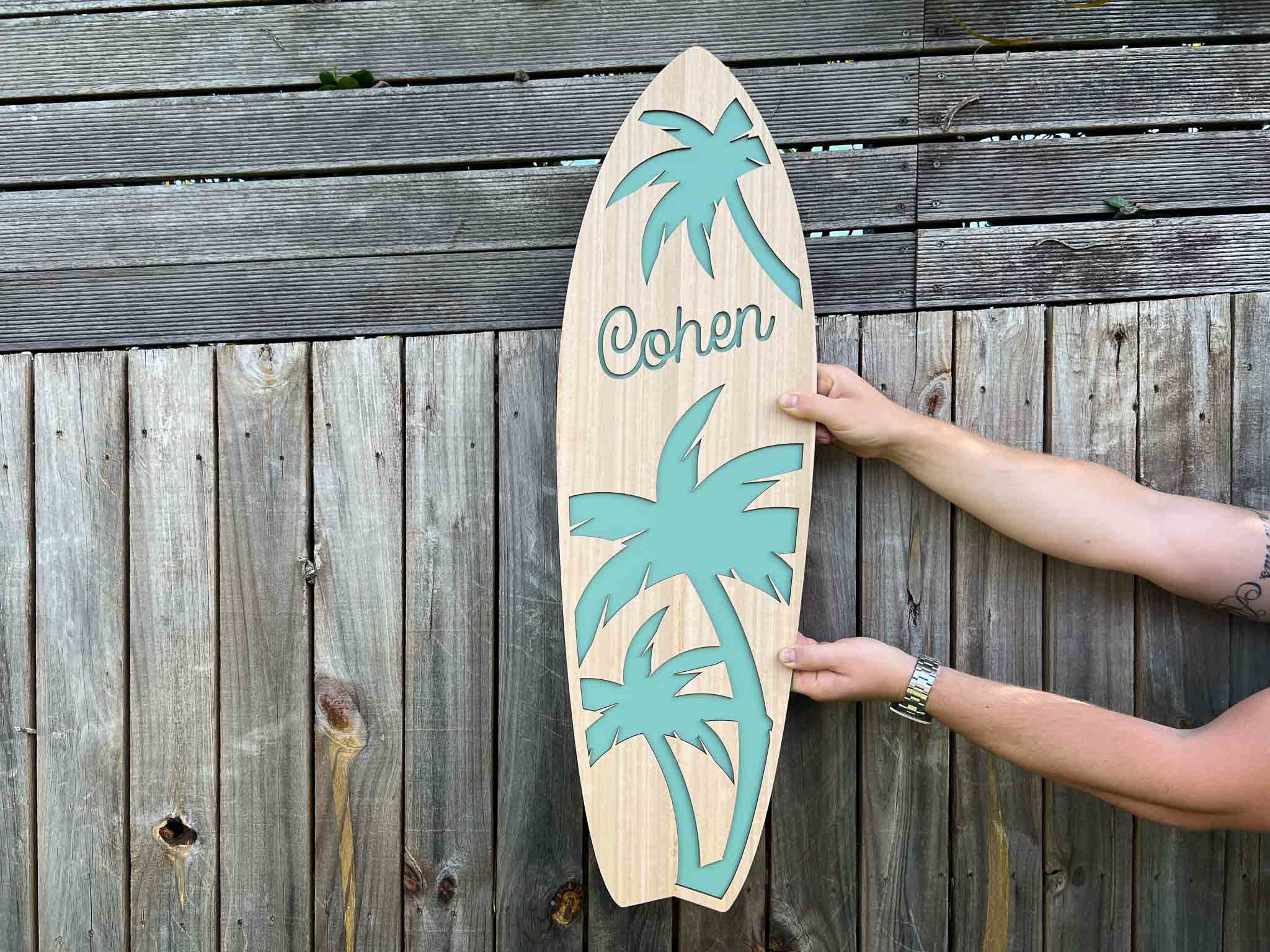 Wooden surfboard-shaped sign with palm tree designs and the name 'Cohen' against a wooden background.