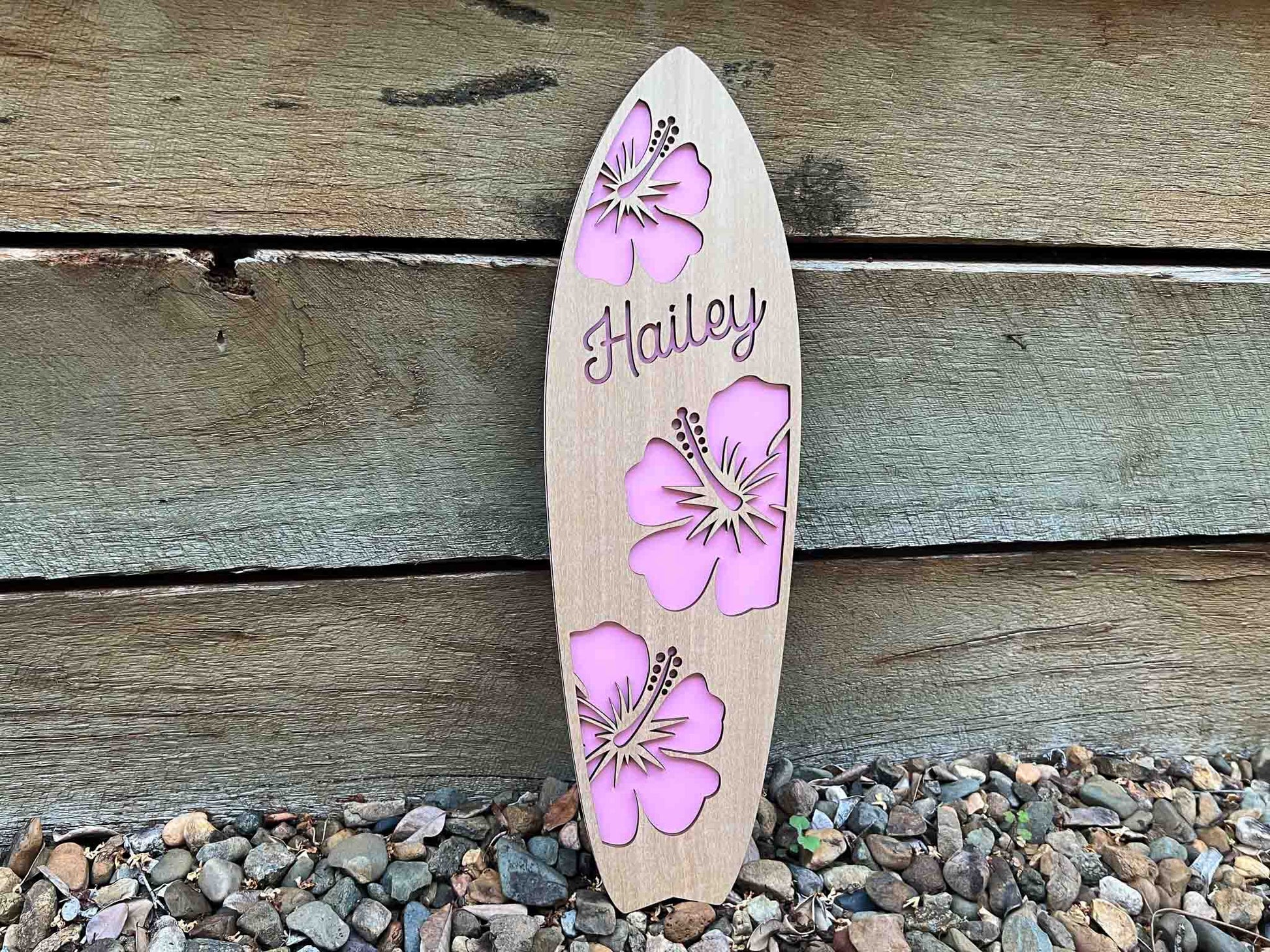 Wooden surfboard-shaped sign with pink floral designs and 'Hailey' text, leaning against a wooden wall.