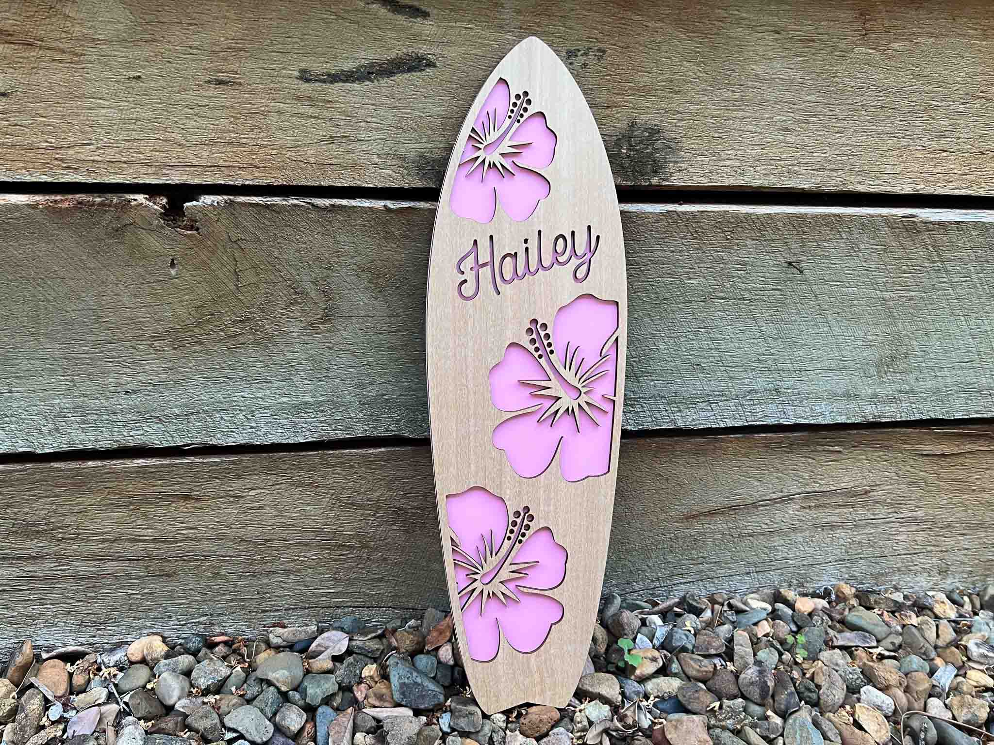 Wooden surfboard-shaped sign with pink floral designs and 'Hailey' text, leaning against a wooden wall.