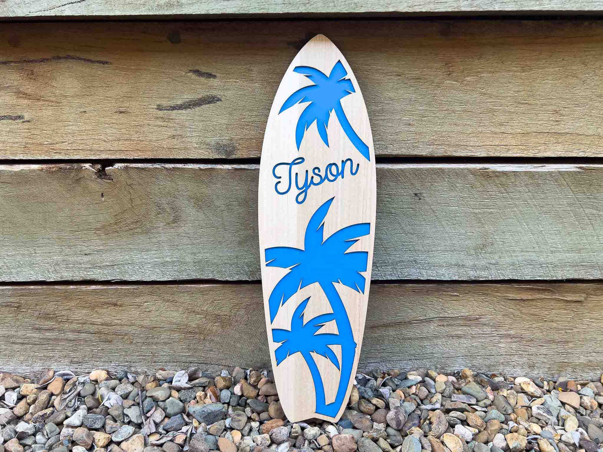 Wooden surfboard with blue palm tree designs and 'Tyson' branding against a wooden background.