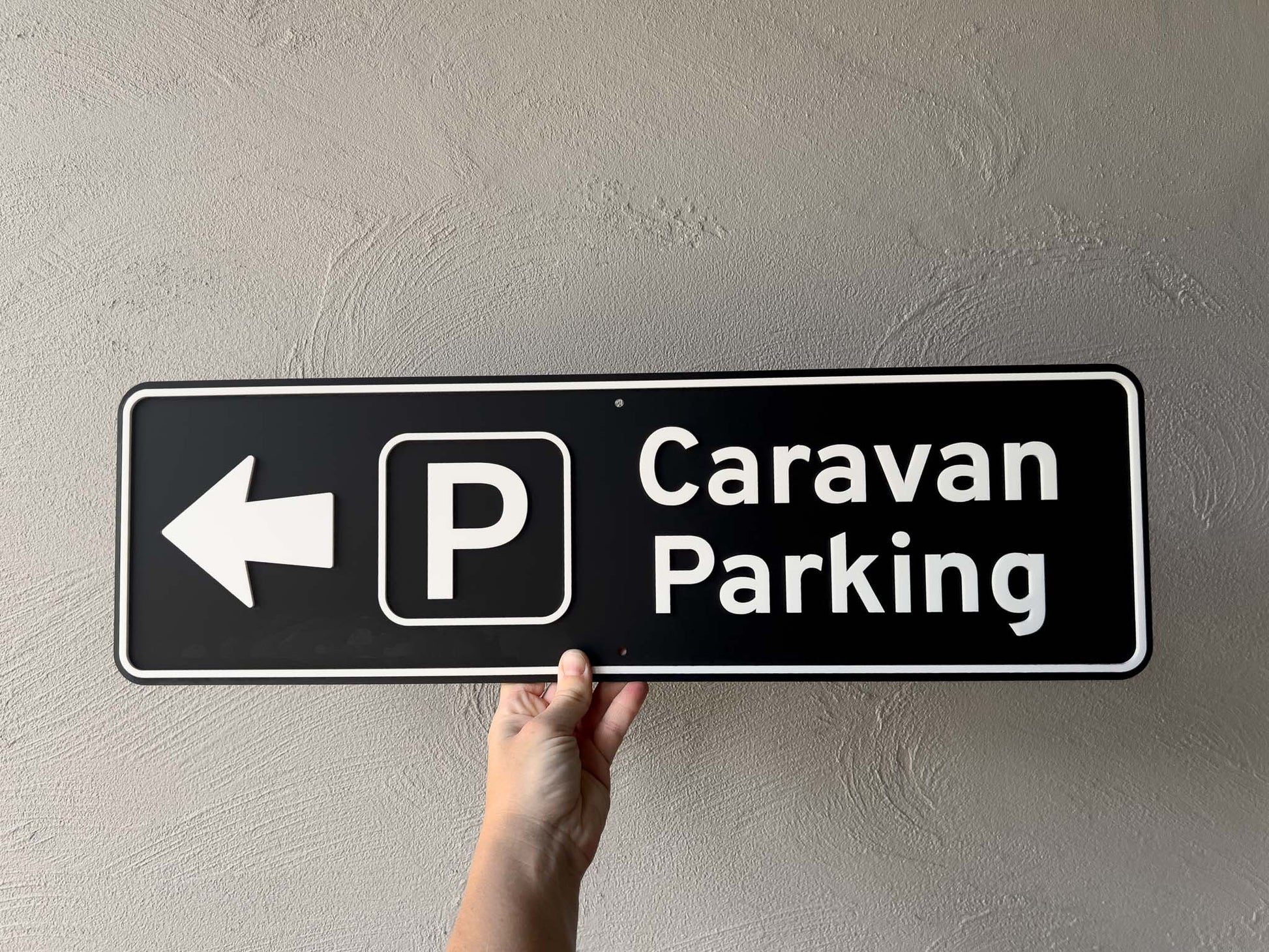 Hand holding a 'Caravan Parking' sign with an arrow on a plain background | Coji Designs