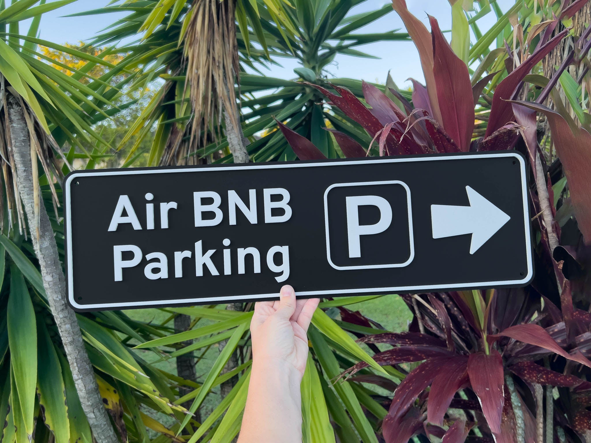 Hand holding a sign that reads 'Air BNB Parking' with a white arrow pointing right against a tropical background | Coji Designs