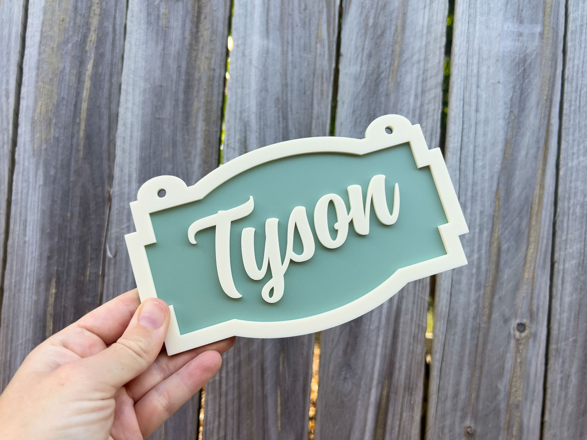 Hand holding a decorative sign with 'Tyson' on it against a wooden fence background | Coji Designs