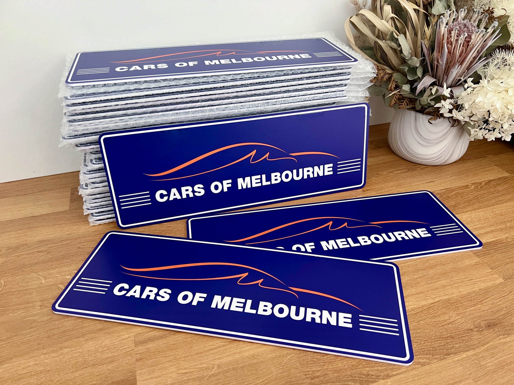 Stack of 'Cars of Melbourne' branded arylic logo plates on a wooden surface with a decorative plant in the background.