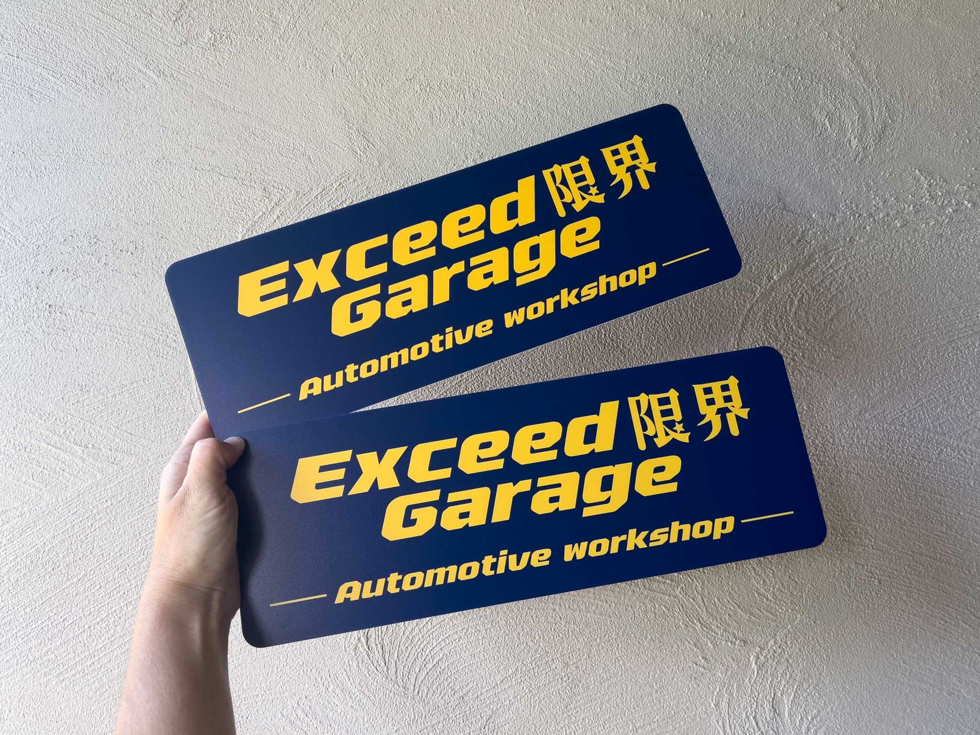 Two 'Exceed Garage' automotive workshop signs held against a light wall.