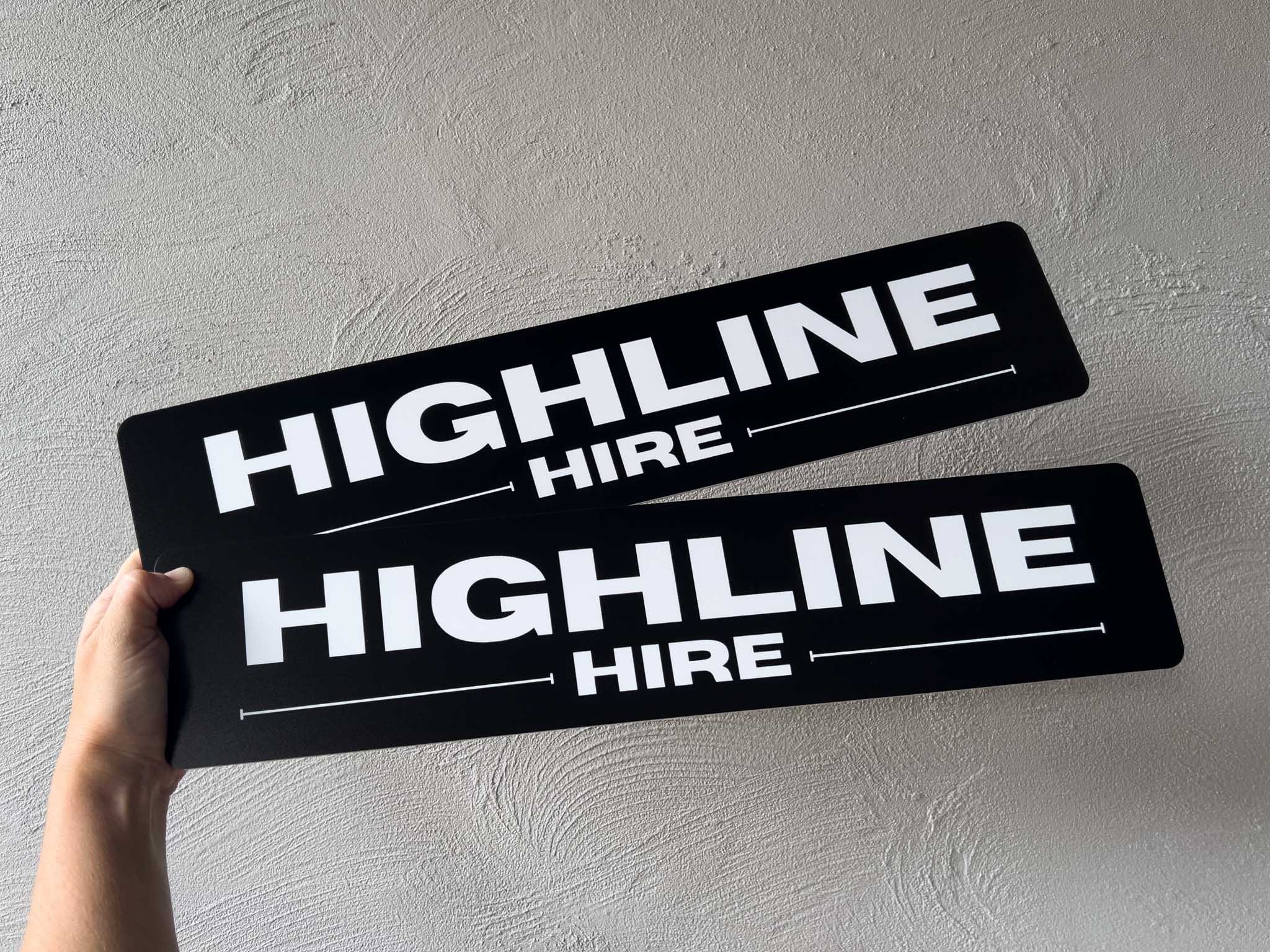 Two black Euro logo plate signs with 'HIGHLINE HIRE' text held against a light gray wall.