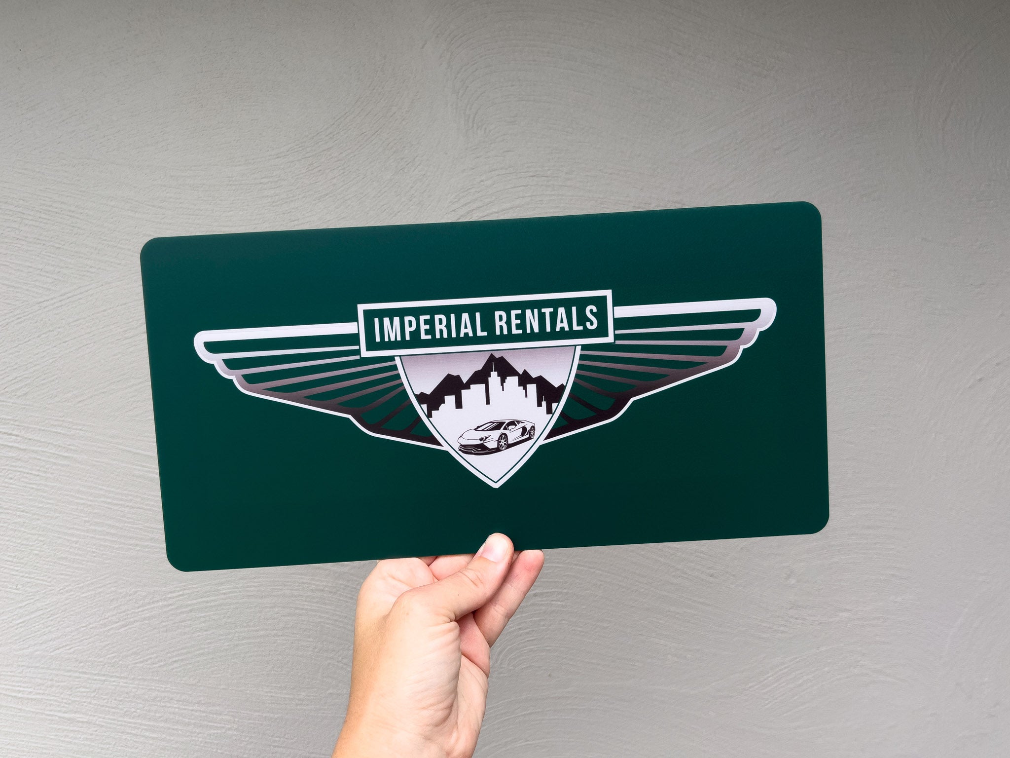 Hand holding a green acrylic plate sign with 'Imperial Rentals' logo against a grey background