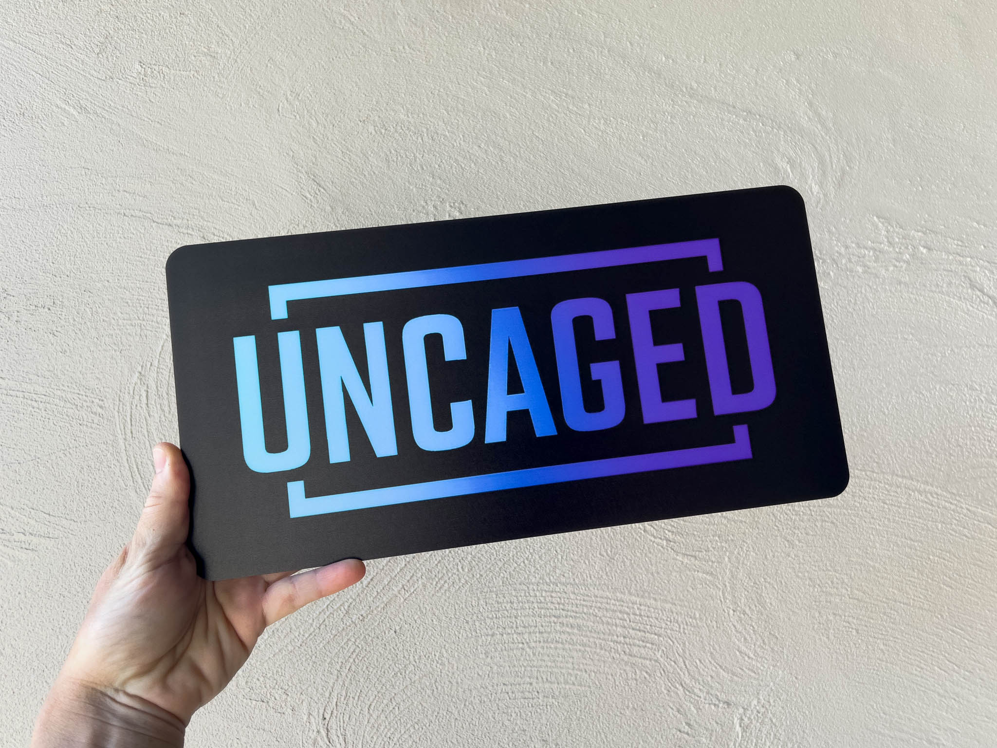 Hand holding a black sign with 'UNCAGED' in blue and purple text against a light grey background