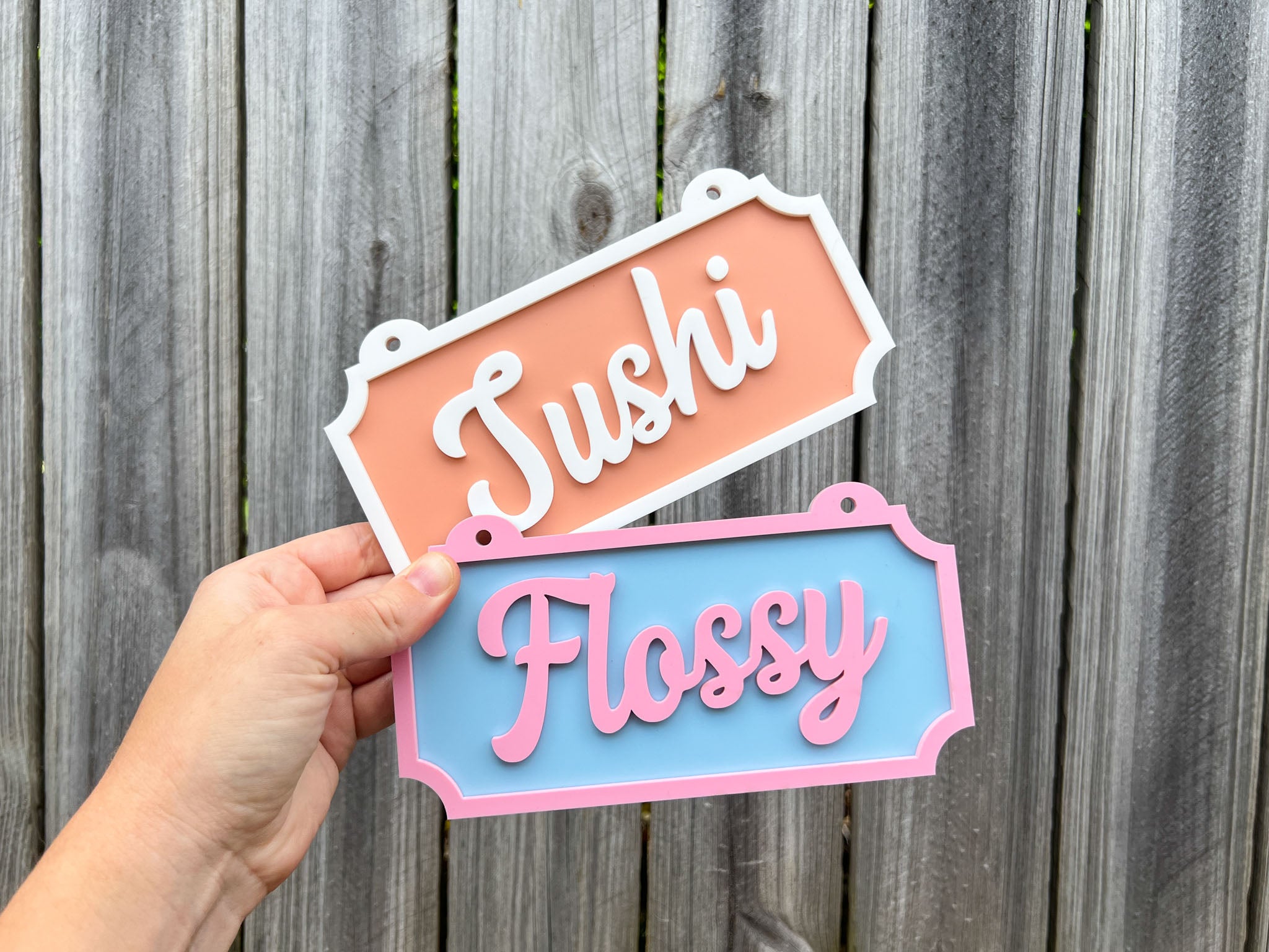 Hand holding two decorative signs with 'Sushi' and 'Flossy' text against a wooden background | Coji Designs