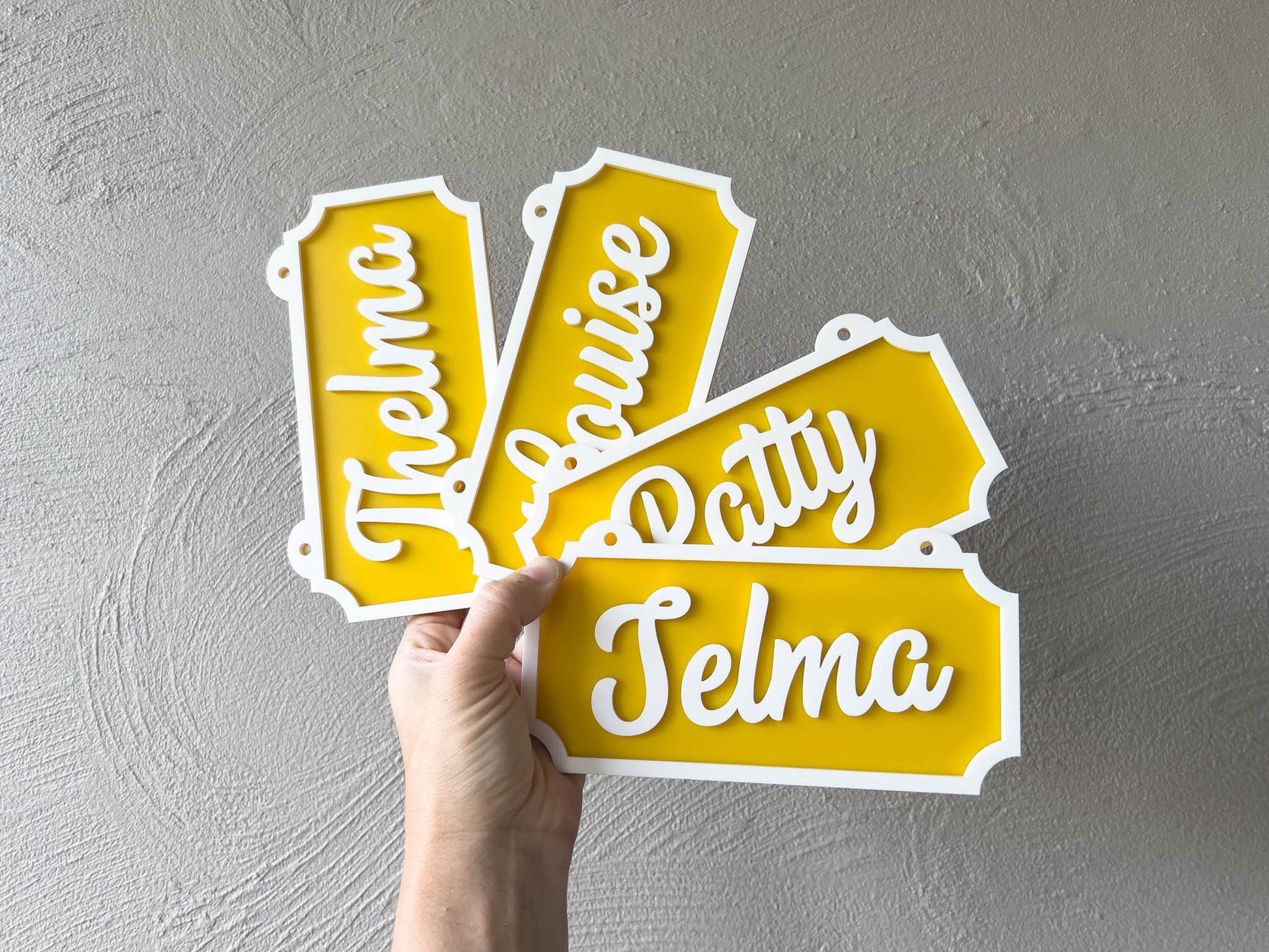 Hand holding yellow name signs with white text against a gray wall | Coji Designs