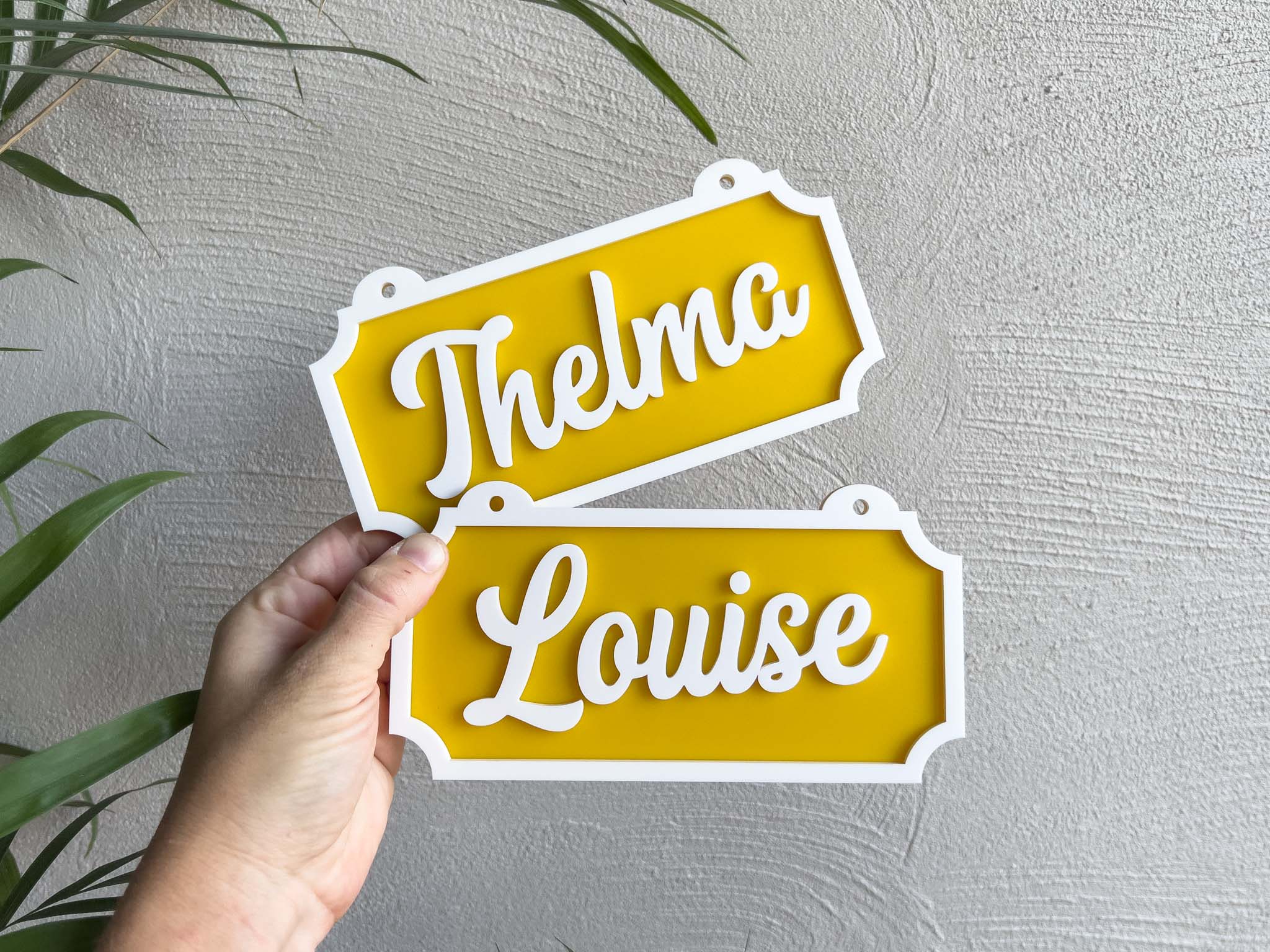 Two yellow signs with 'Thelma' and 'Louise' text held against a light grey wall | Coji Designs