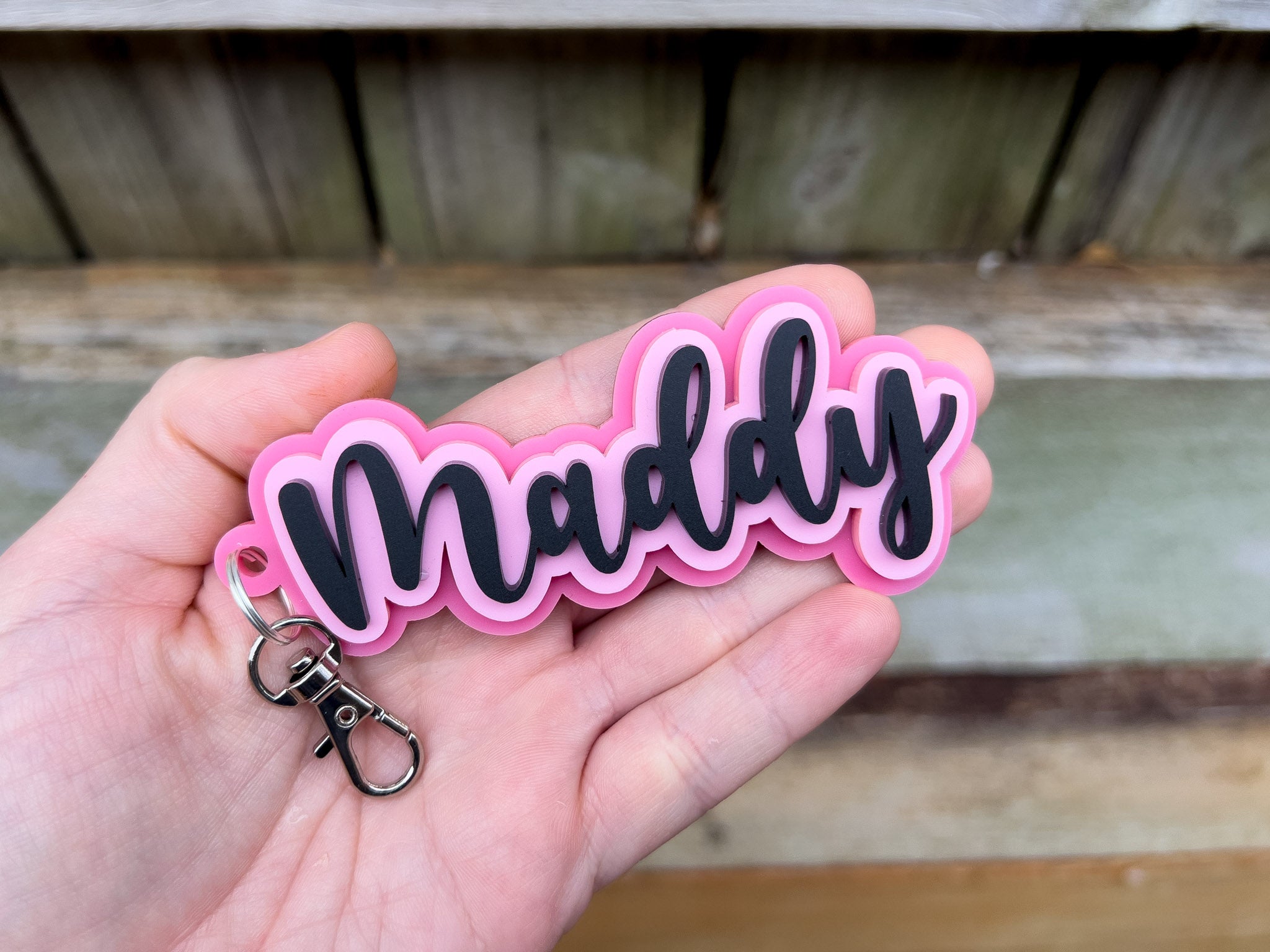Keychain with 'Maddy' in pink and black held in a hand against a wooden background