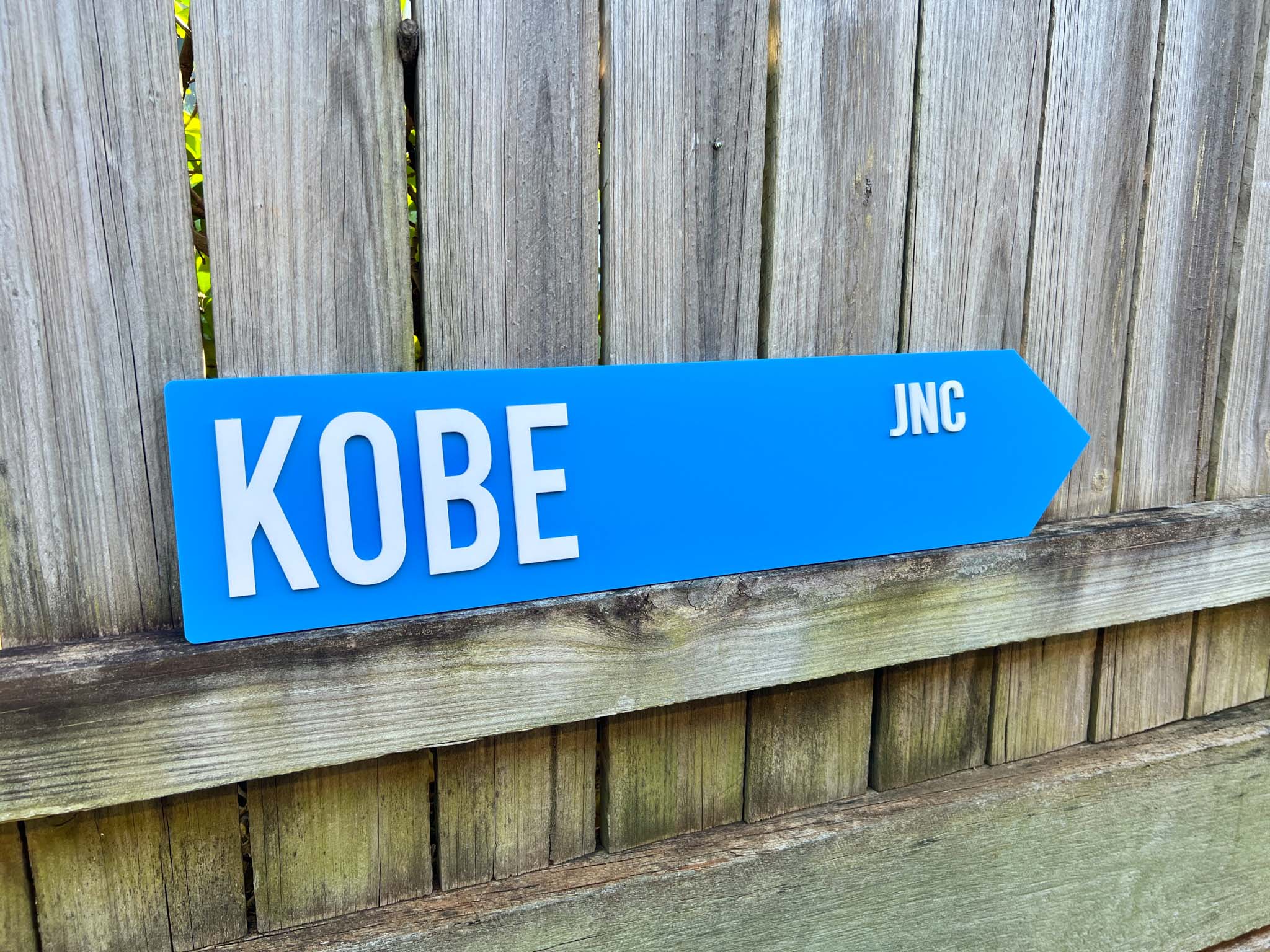 Blue sign pointing to 'Kobe' with 'JNC' on a wooden fence background | Coji Designs