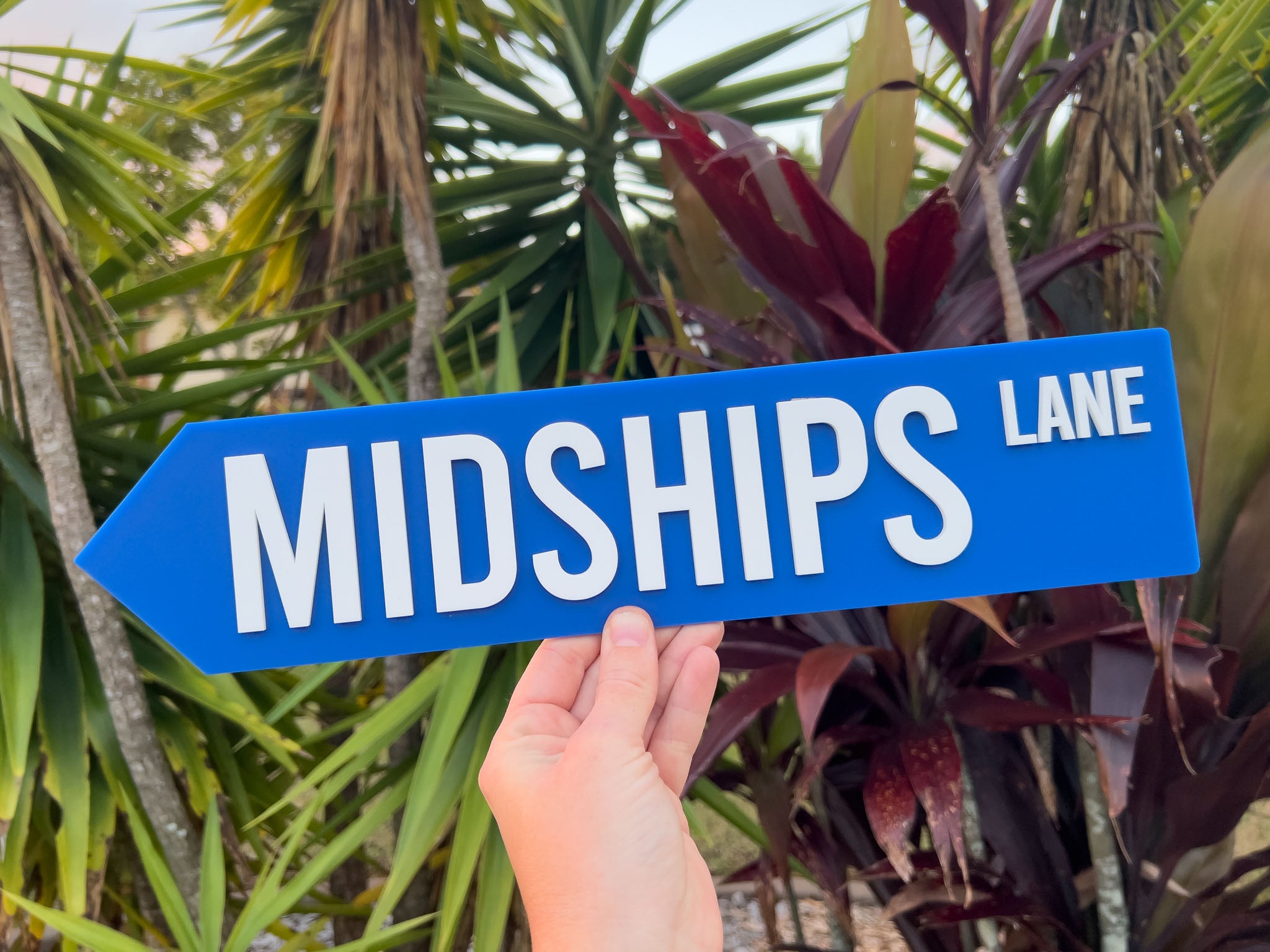 Hand holding a blue sign with 'MIDSHIPS LANE' text against a tropical plant background | Coji Designs
