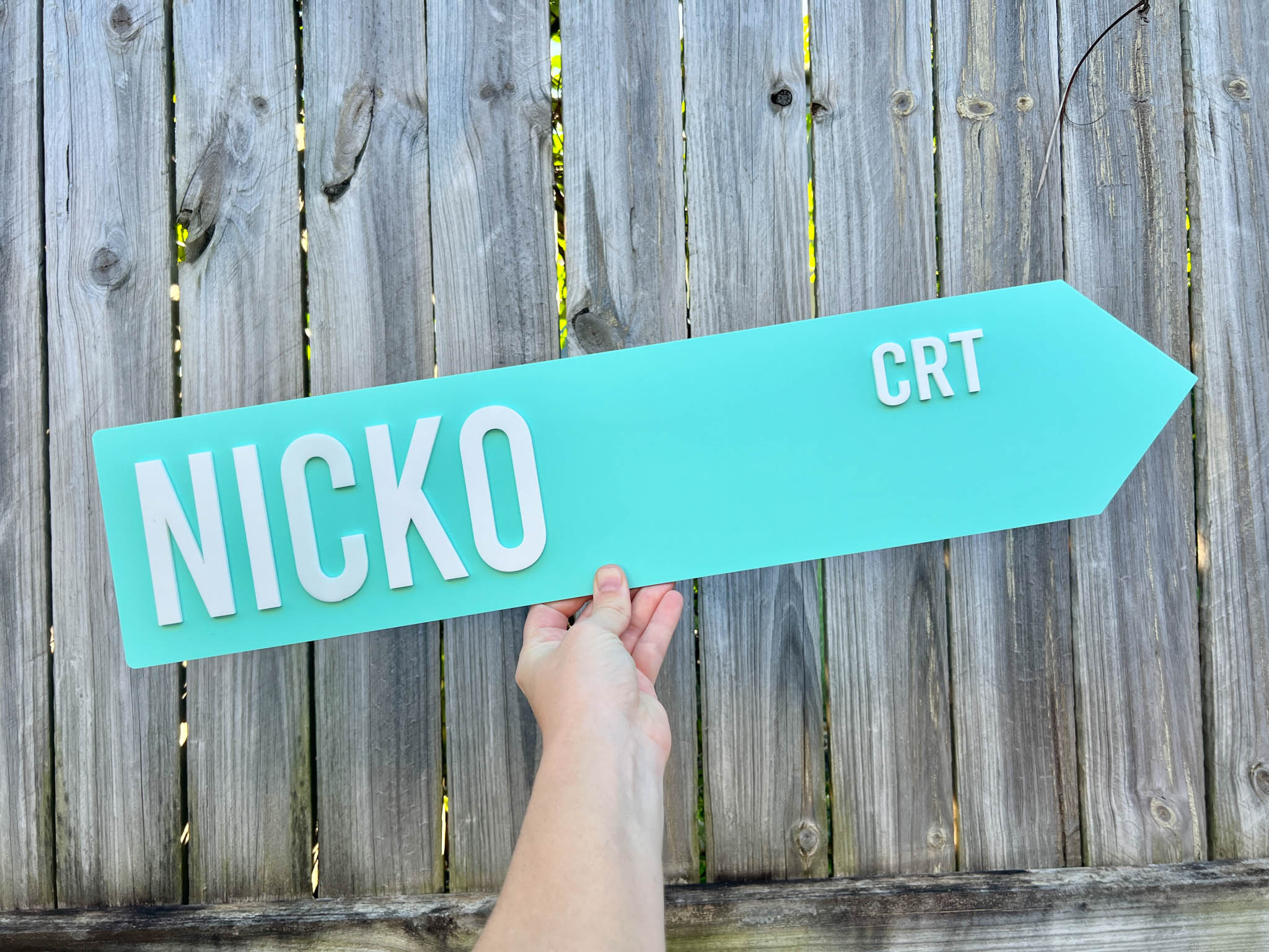 Hand holding a teal arrow-shaped sign with 'NICKO CRT' text against a wooden fence background | Coji Designs