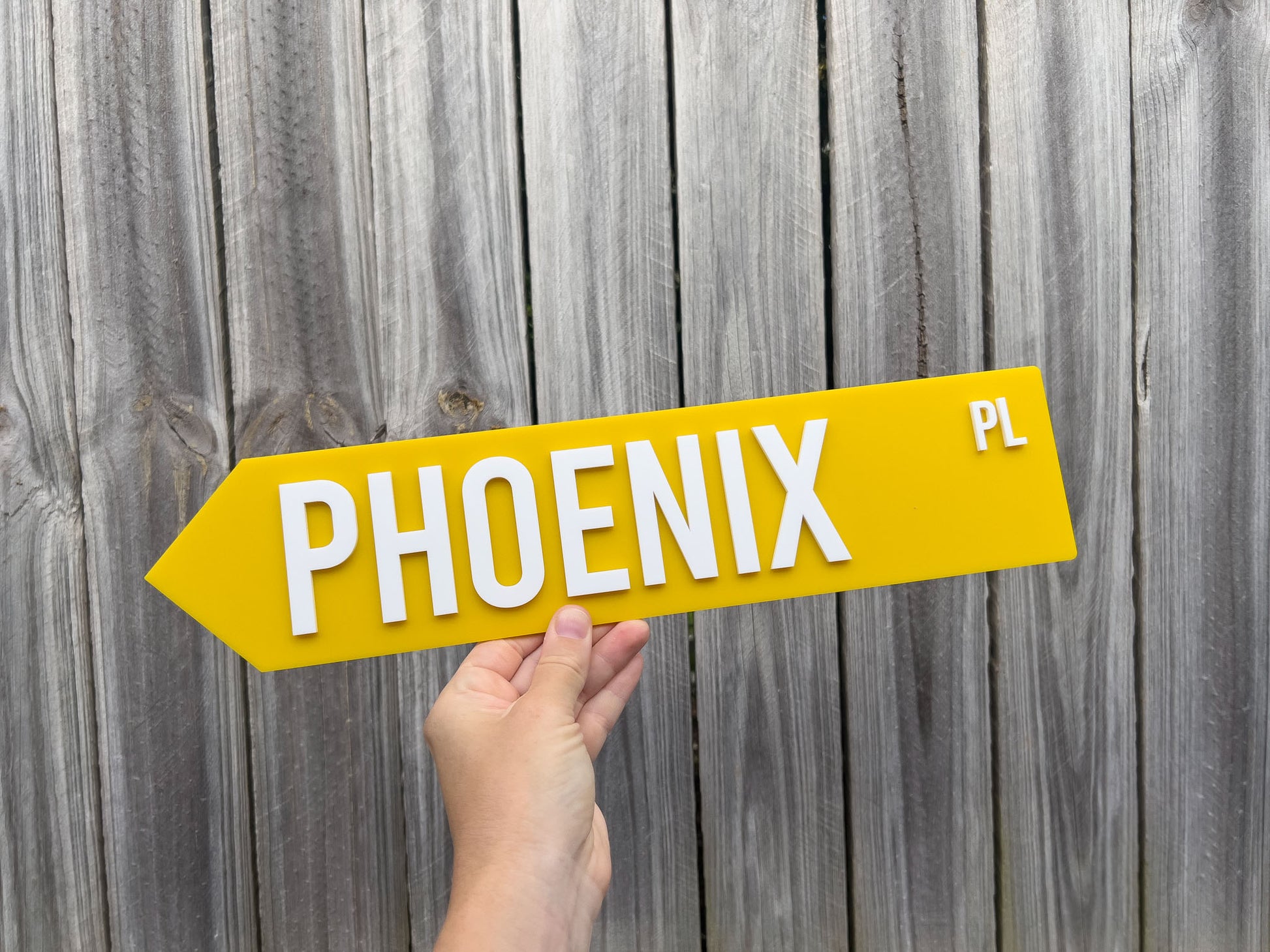 Hand holding a yellow street sign with 'PHOENIX PL' against a wooden fence background | Coji Designs