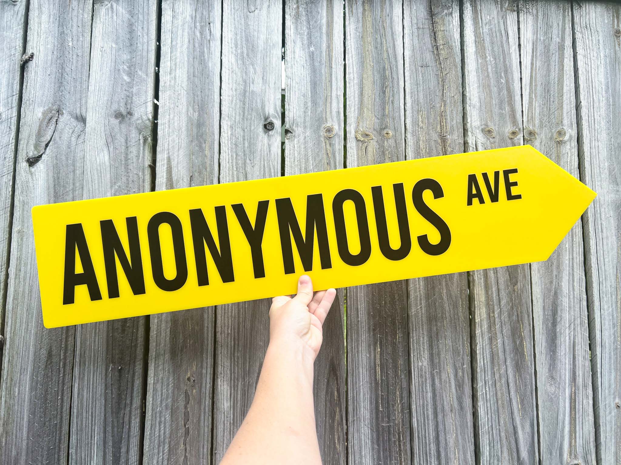 Hand holding a yellow sign with 'ANONYMOUS AVE' text against a wooden background