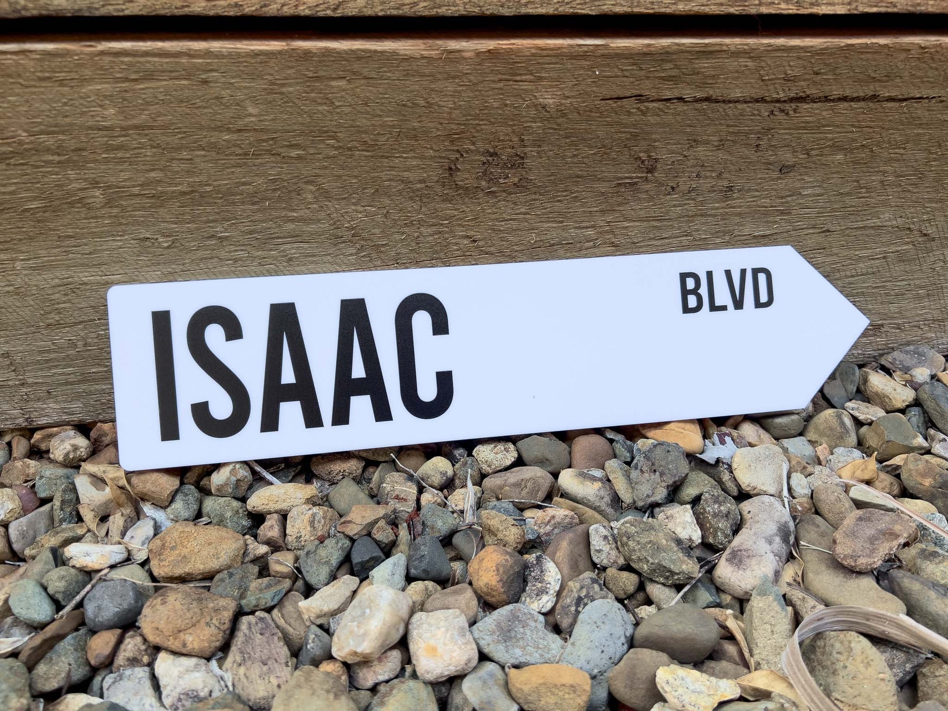 White street sign with 'ISAAC BLVD' text on a wooden surface and pebbles below