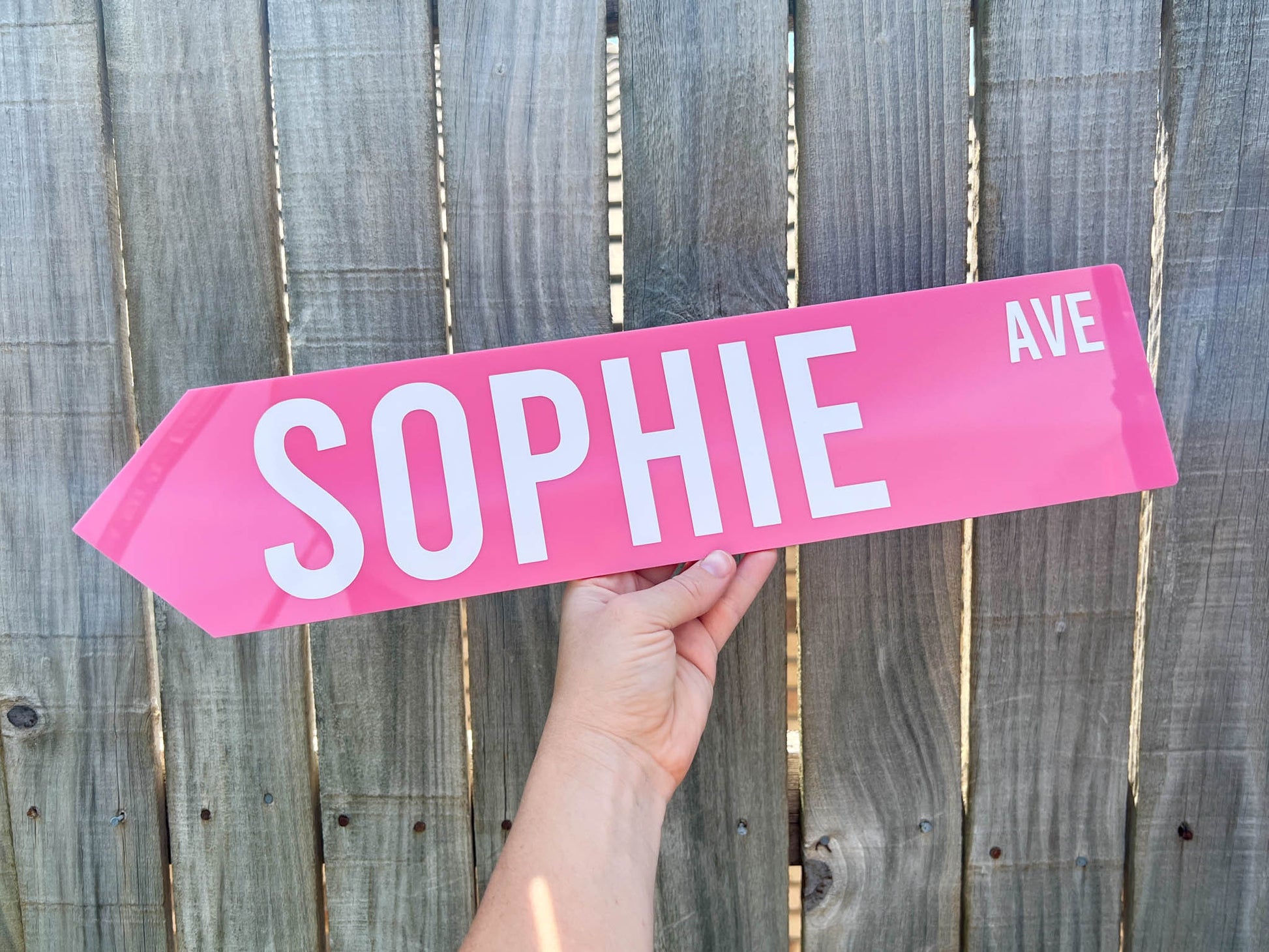 Pink street sign with 'SOPHIE AVE' held in front of a wooden fence