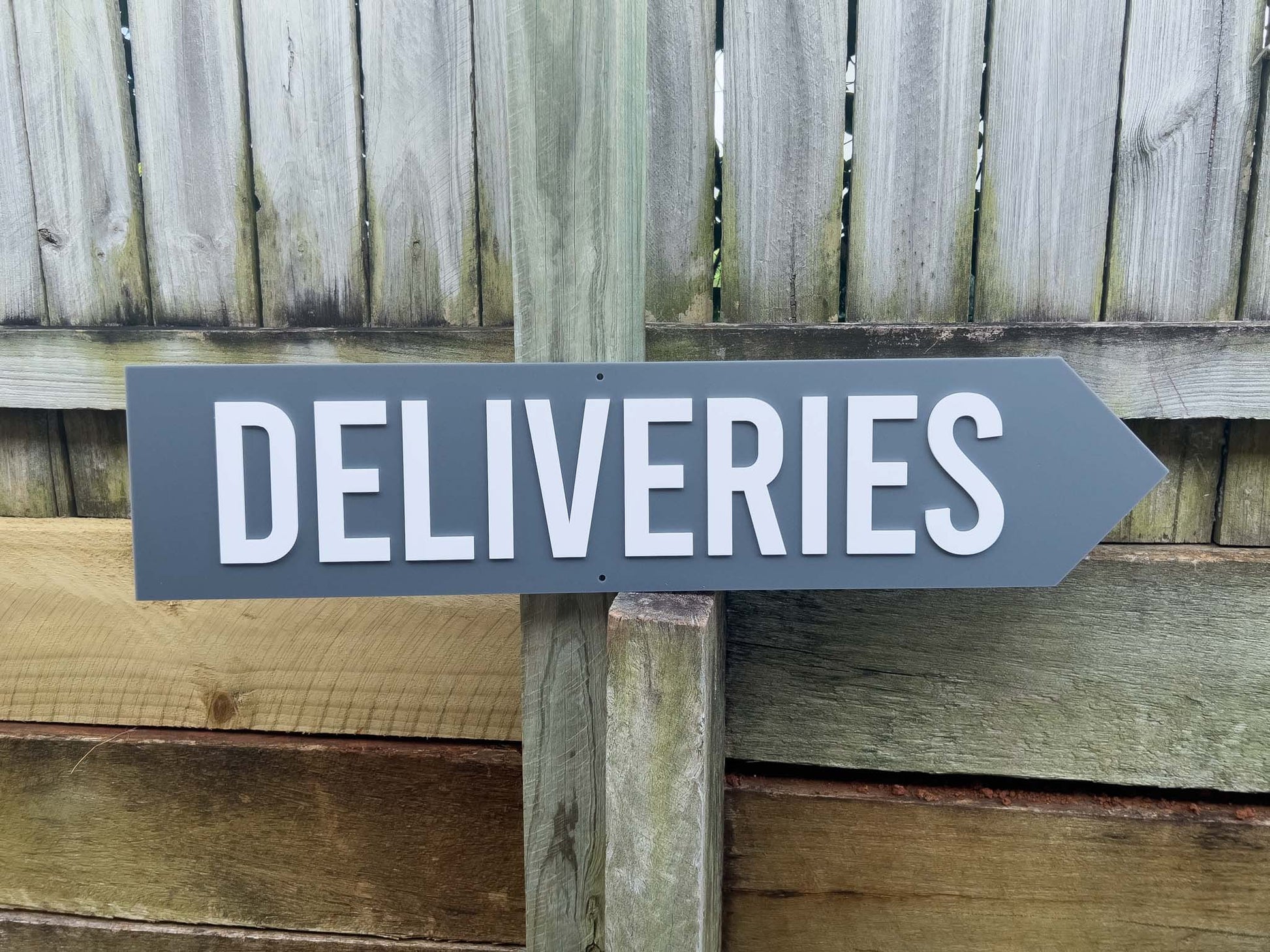 Grey sign with 'DELIVERIES' text and arrow pointing to the right against a wooden fence background | Coji Designs