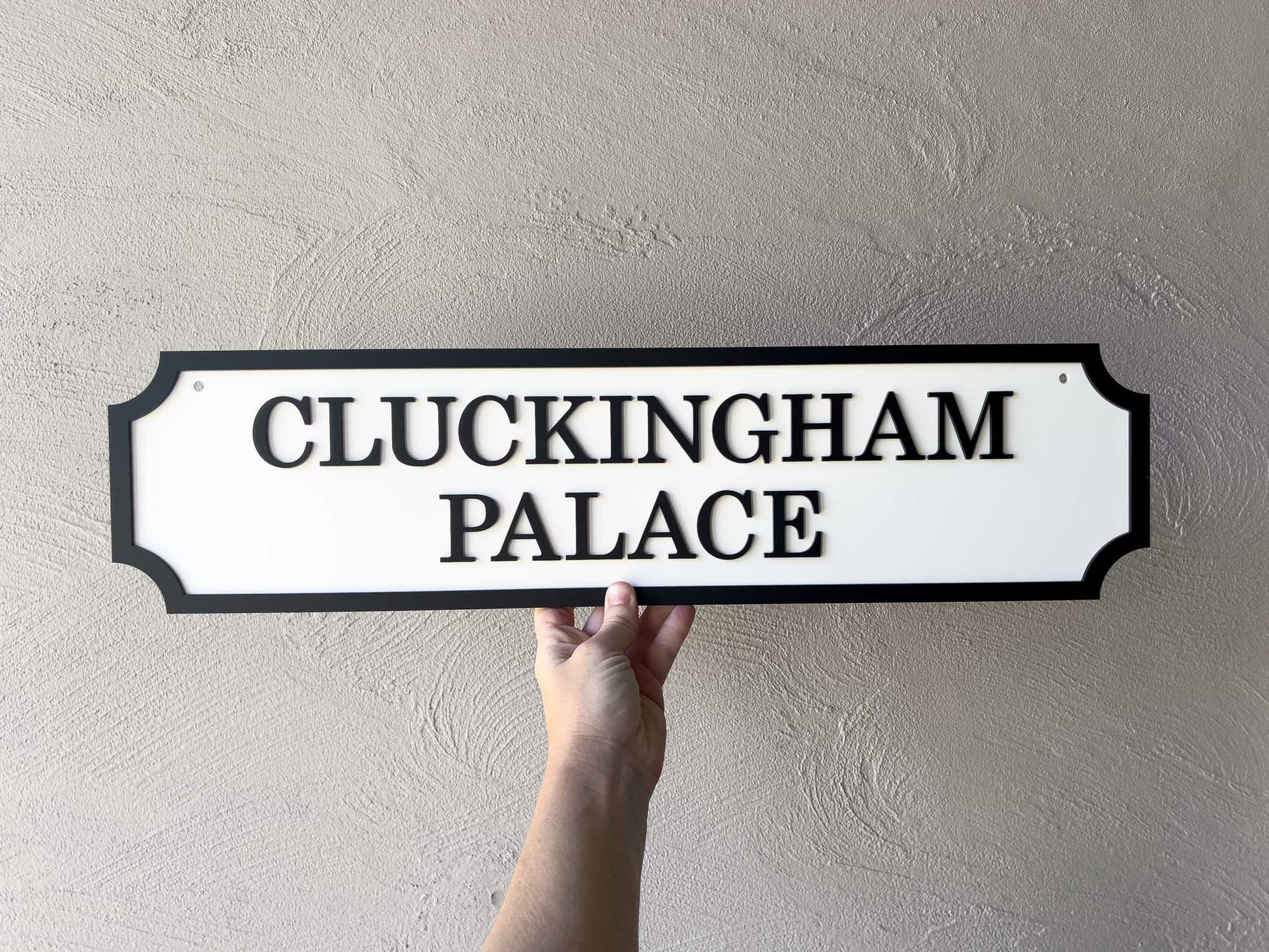 Hand holding a 3D acrylic sign that reads 'Cluckingham Palace' in white and black.