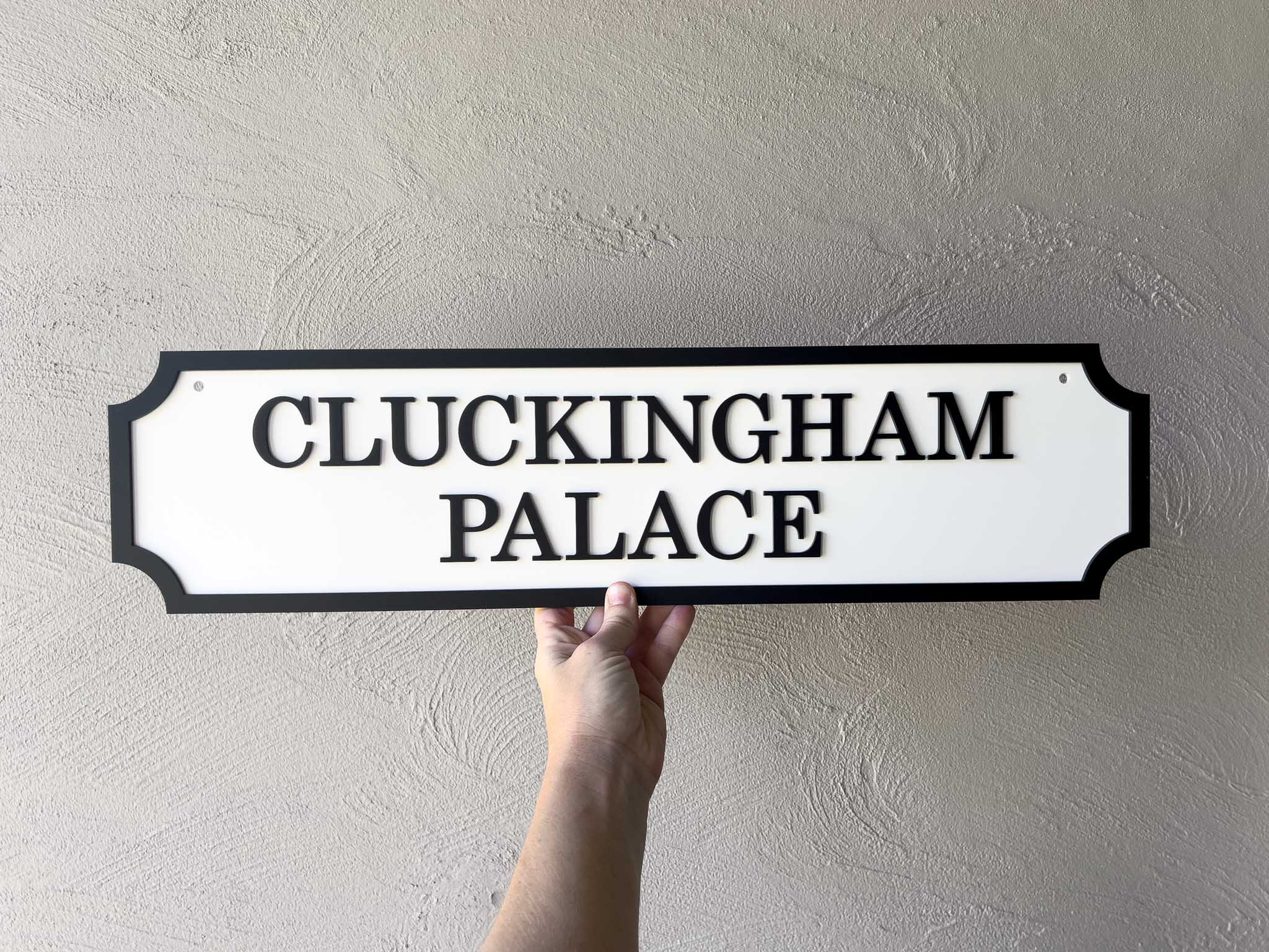 Hand holding a 3D acrylic sign that reads 'Cluckingham Palace' in white and black.