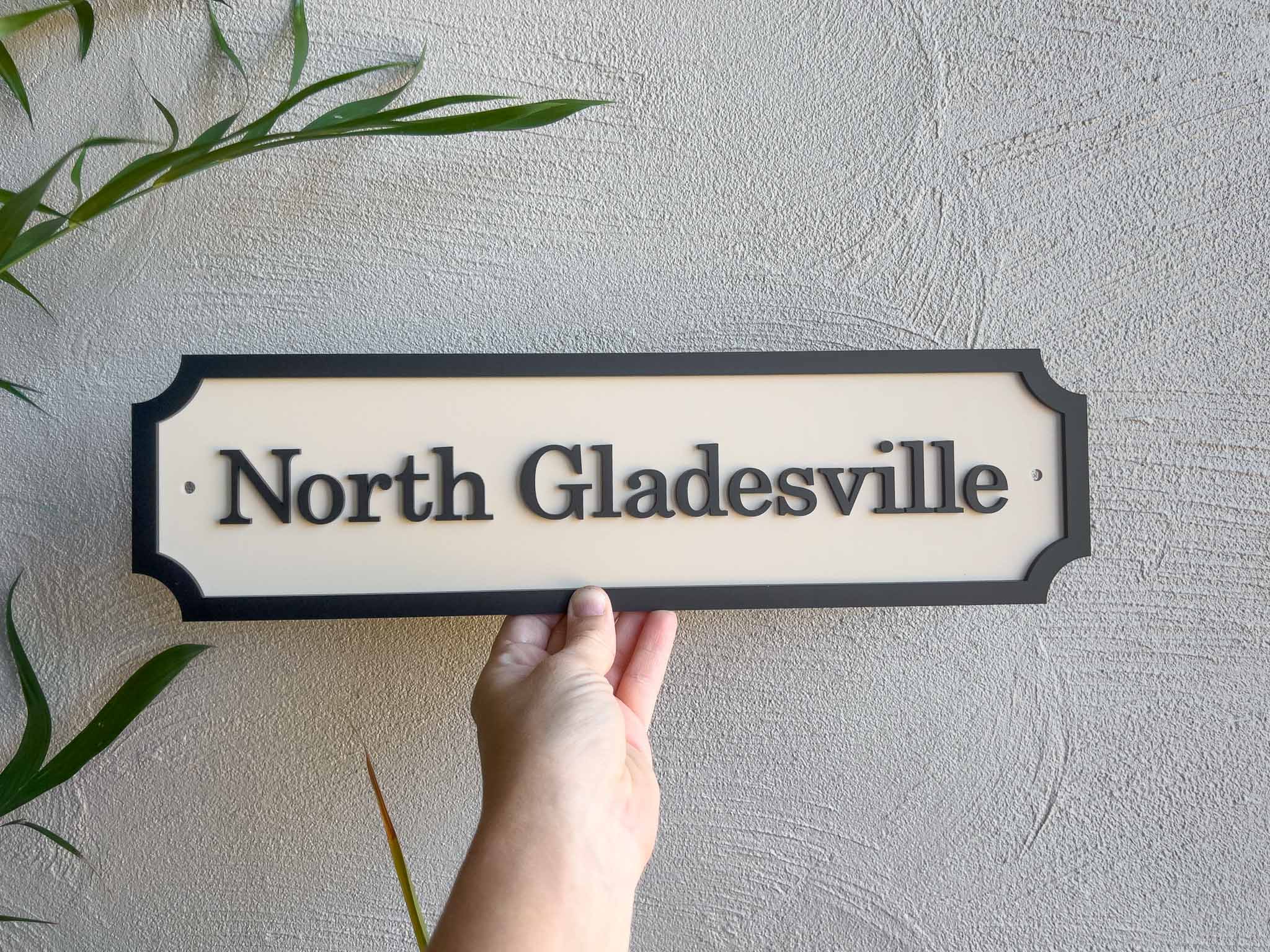 Hand holding a custom made 3D acrylic sign with 'North Gladesville' text against a light gray wall.