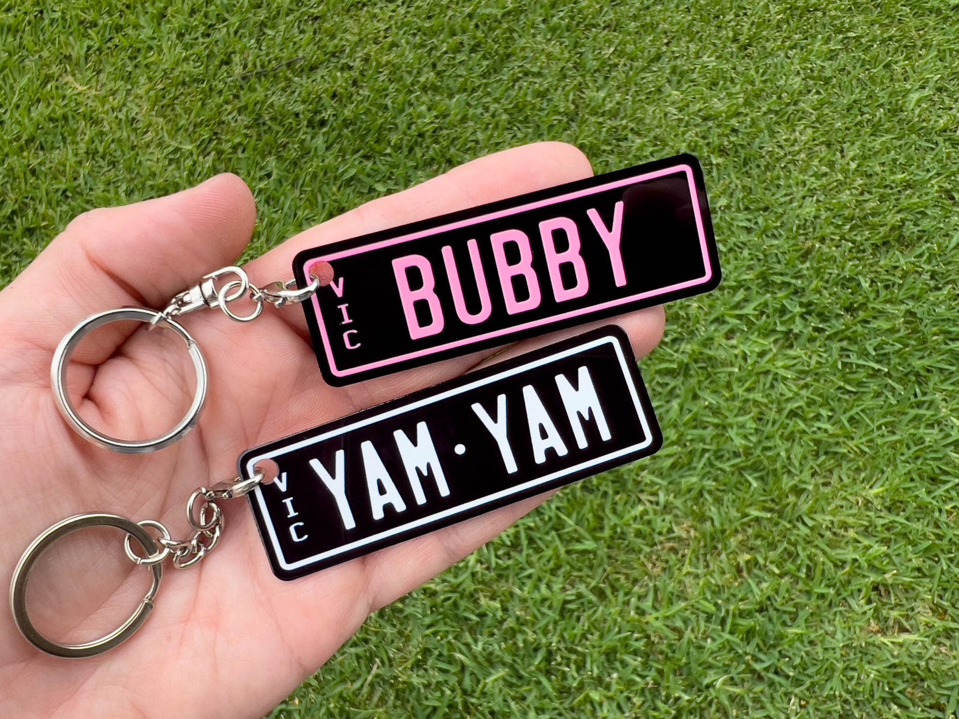 Two keychains with 'BUBBY' and 'YAM YAM' text held in a hand against a grass background.