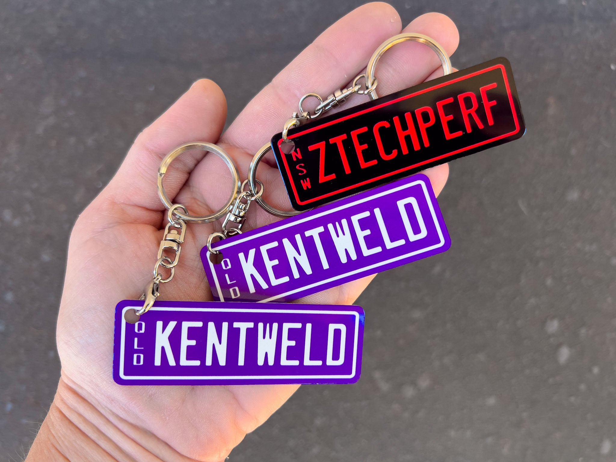 Hand holding three keychains with 'ZTECHPERF', 'KENTWELD', and 'KENTWELD' text on a dark background