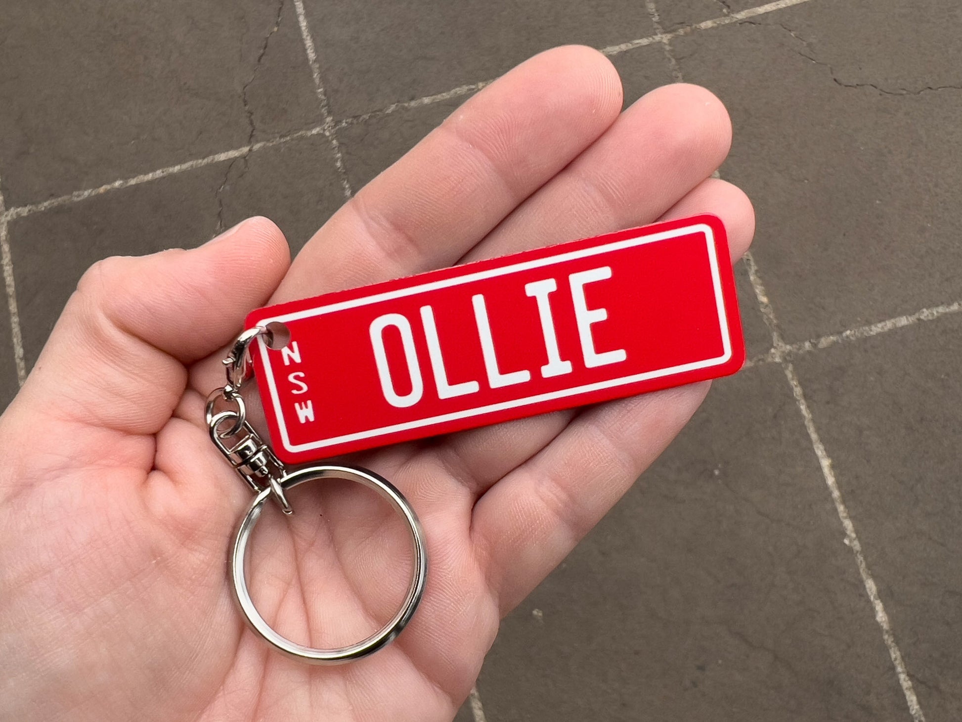 Hand holding a red keychain with 'OLLIE' printed on it against a gray pavement background