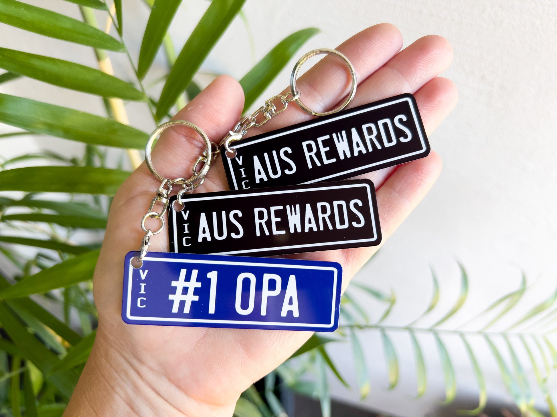 Hand holding three number plate style keychains with 'AUS Rewards' and '#1 OPA' text against a tropical background.