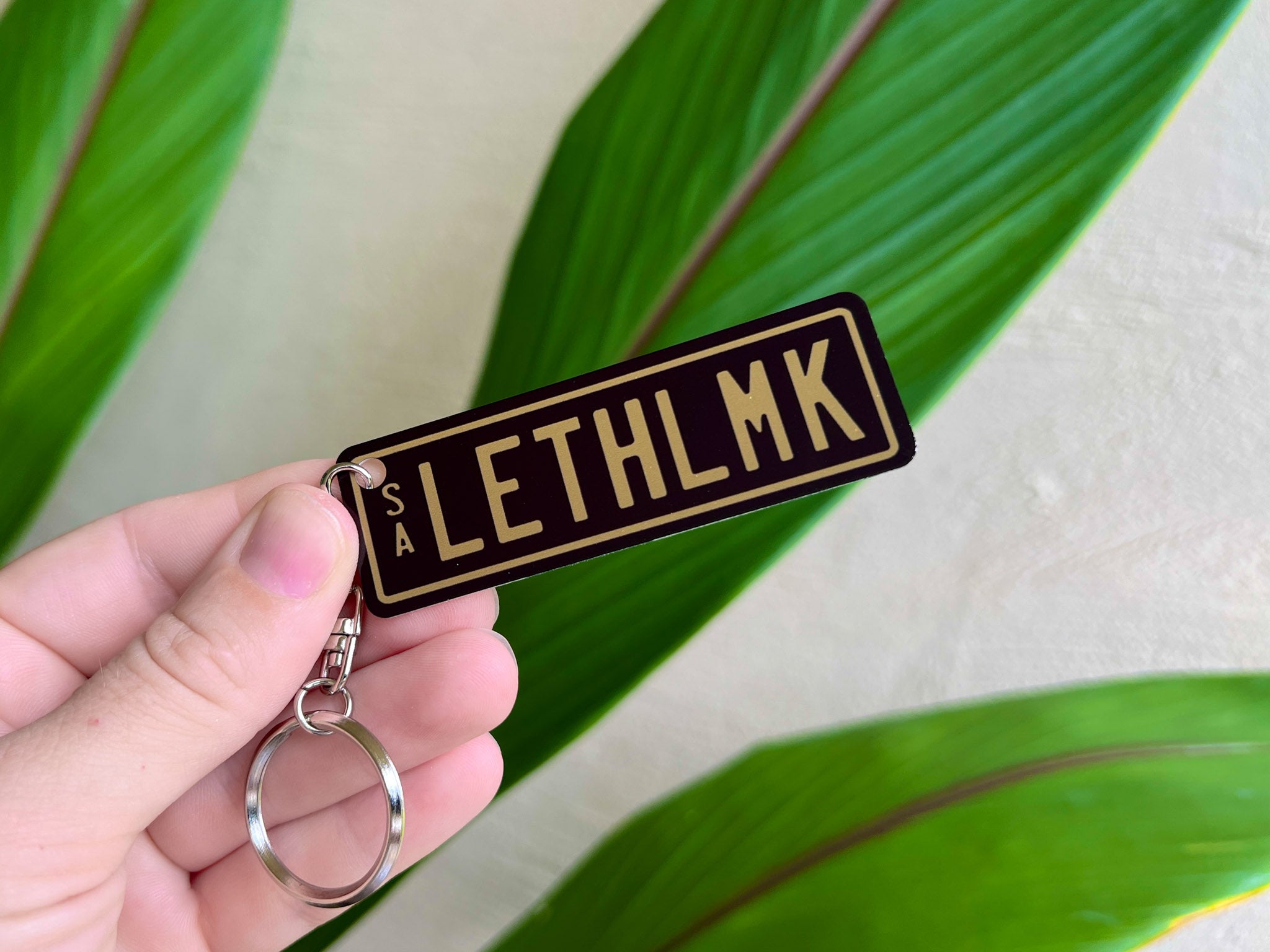 Keychain with 'LETHLMK' text held by a hand against a leafy background