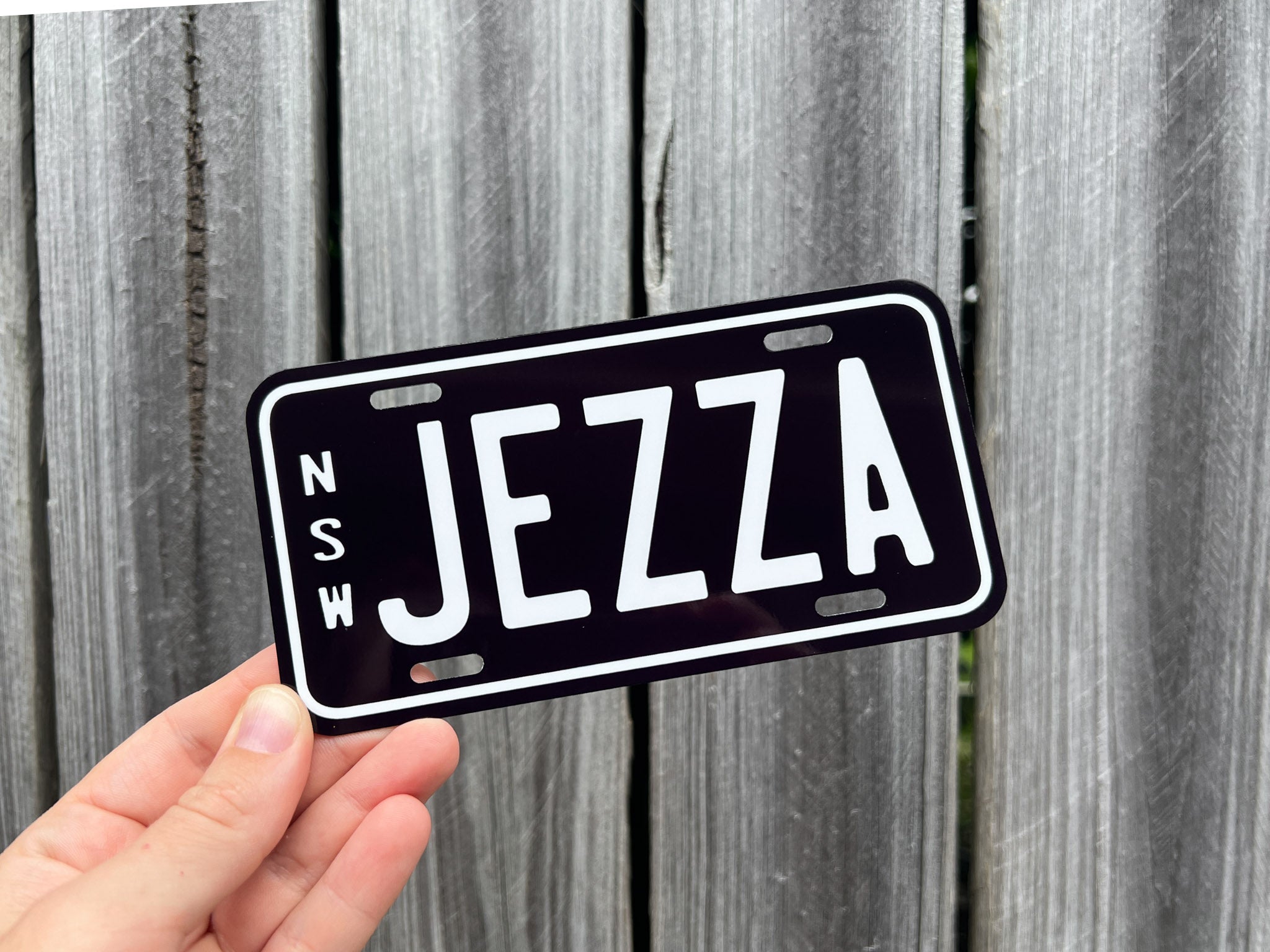 Hand holding a mini black number plate with 'NSW Jezza' text against a wooden background