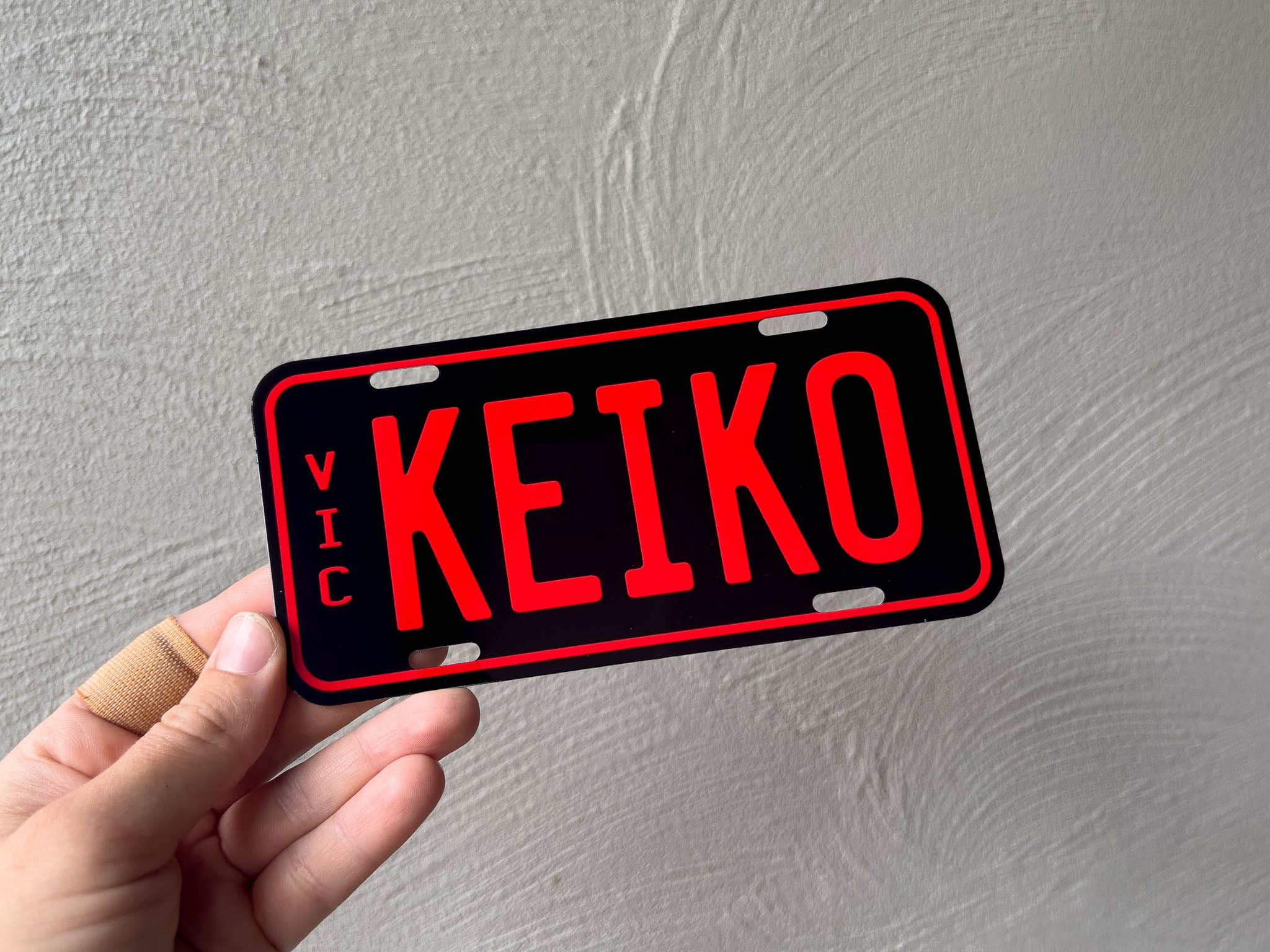Hand holding a red and black mini number plate with 'KEIKO' and 'VIC' text against a grey background