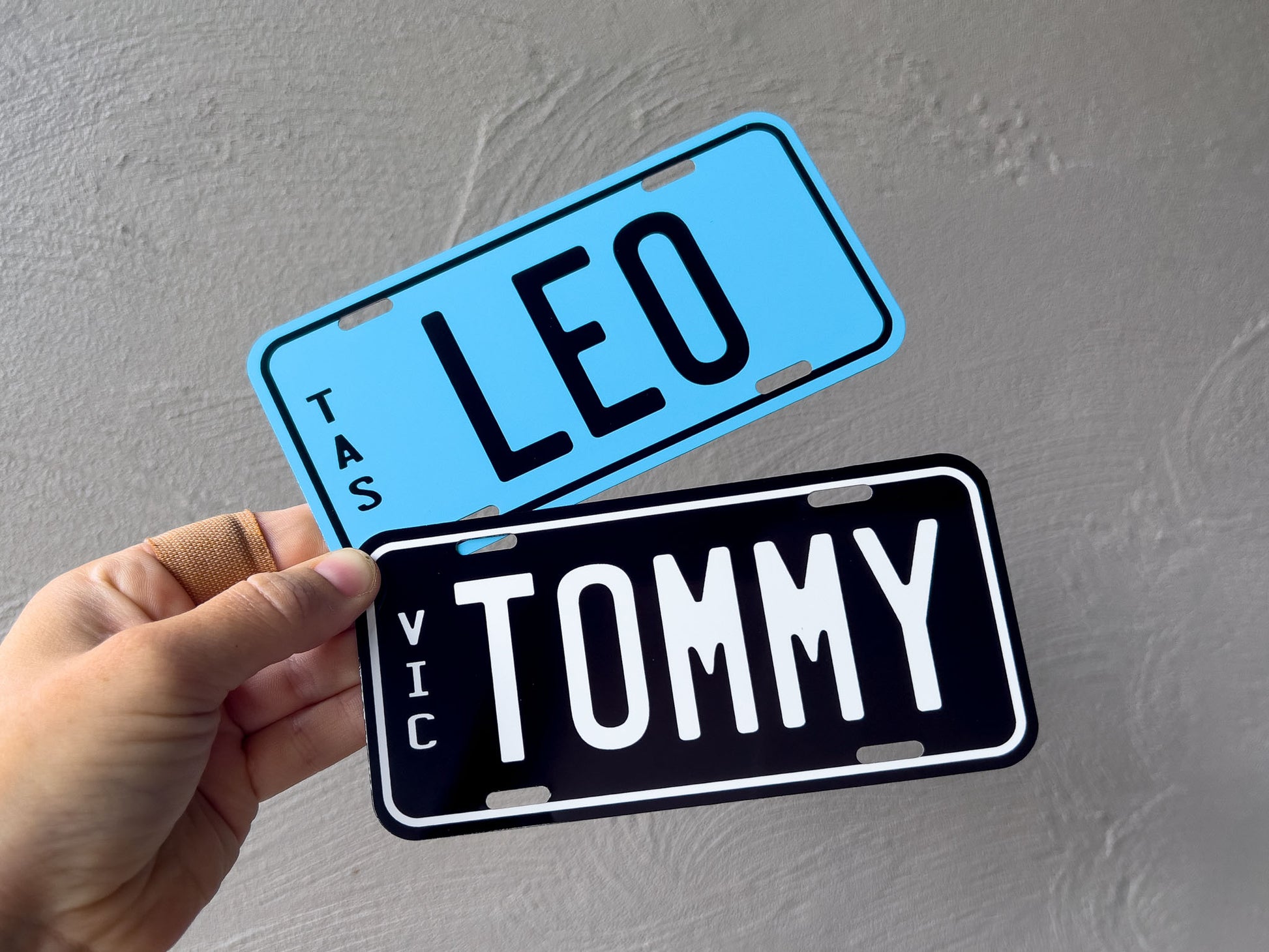Two small number plates, one blue with 'LEO' and another black with 'Tommy', held against a grey background.