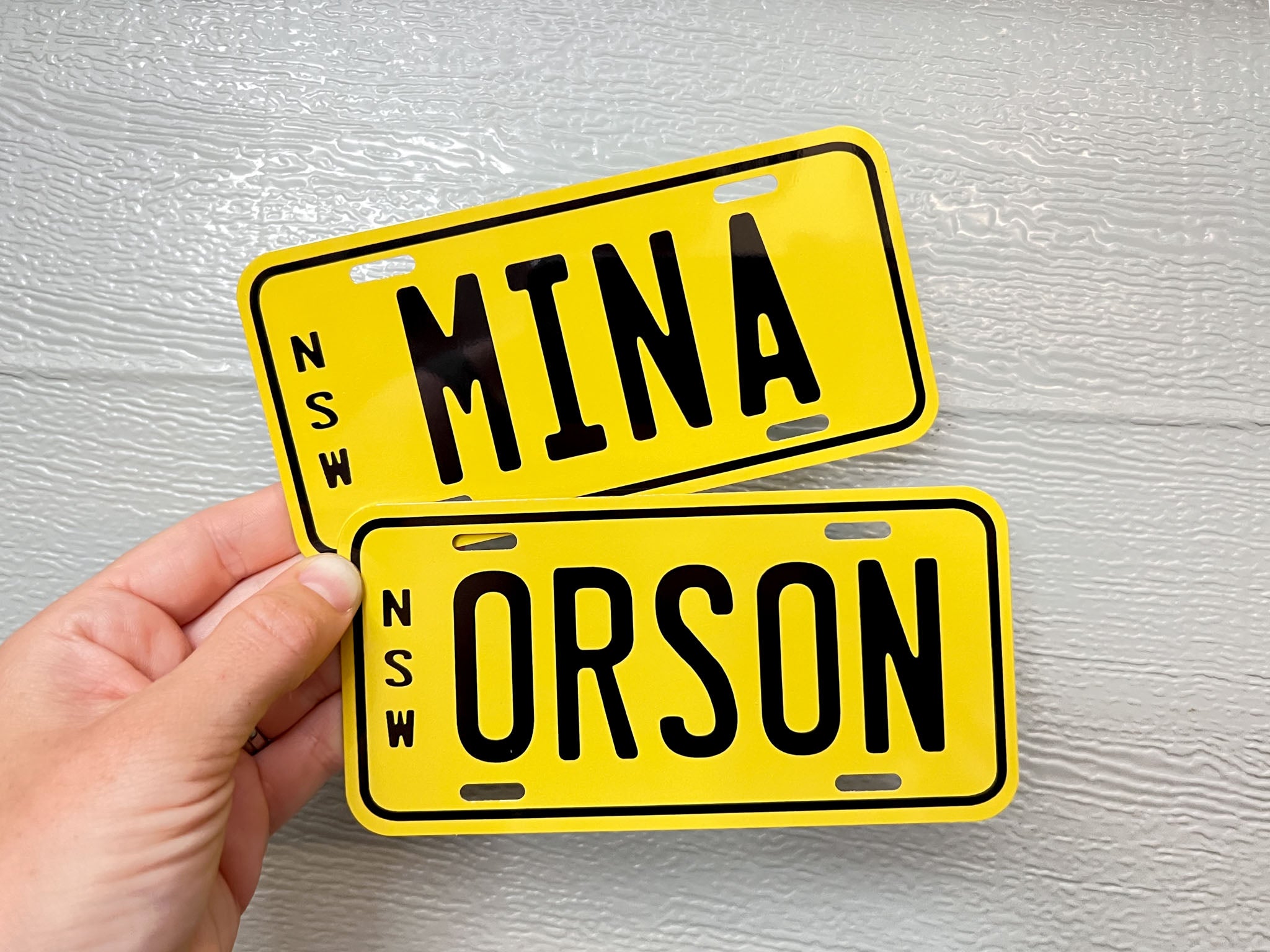 Two yellow mini kid's number plates with black text held by a hand on a grey background