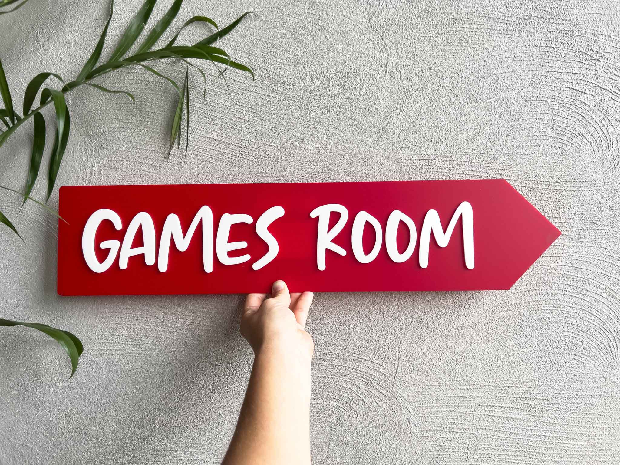 Hand holding a red arrow-shaped sign with 'GAMES ROOM' text against a light gray wall | Coji Designs