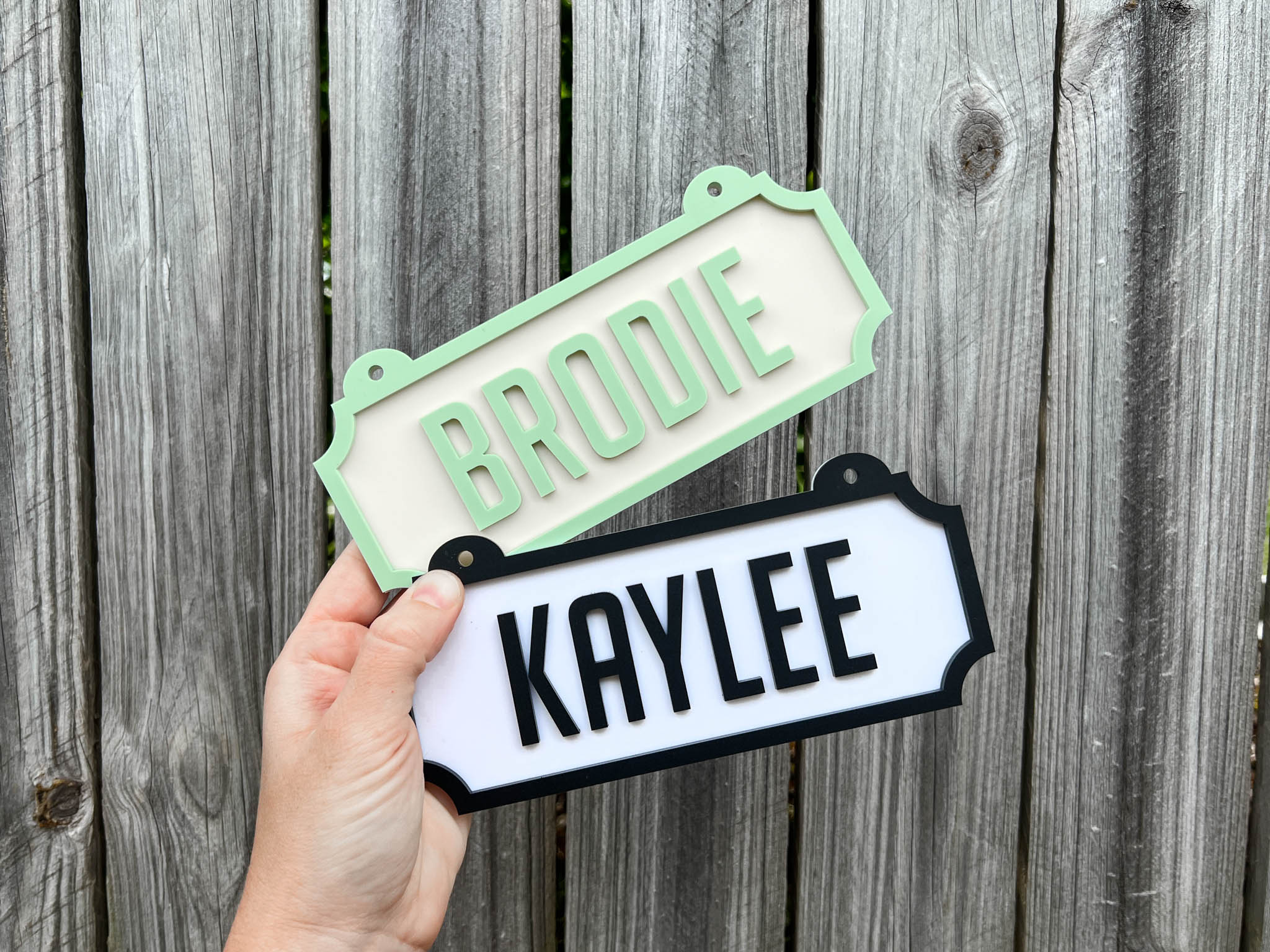 Hand holding two name signs, one green with 'Brodie' and one white with 'Kaylee', against a wooden background  | Coji Designs